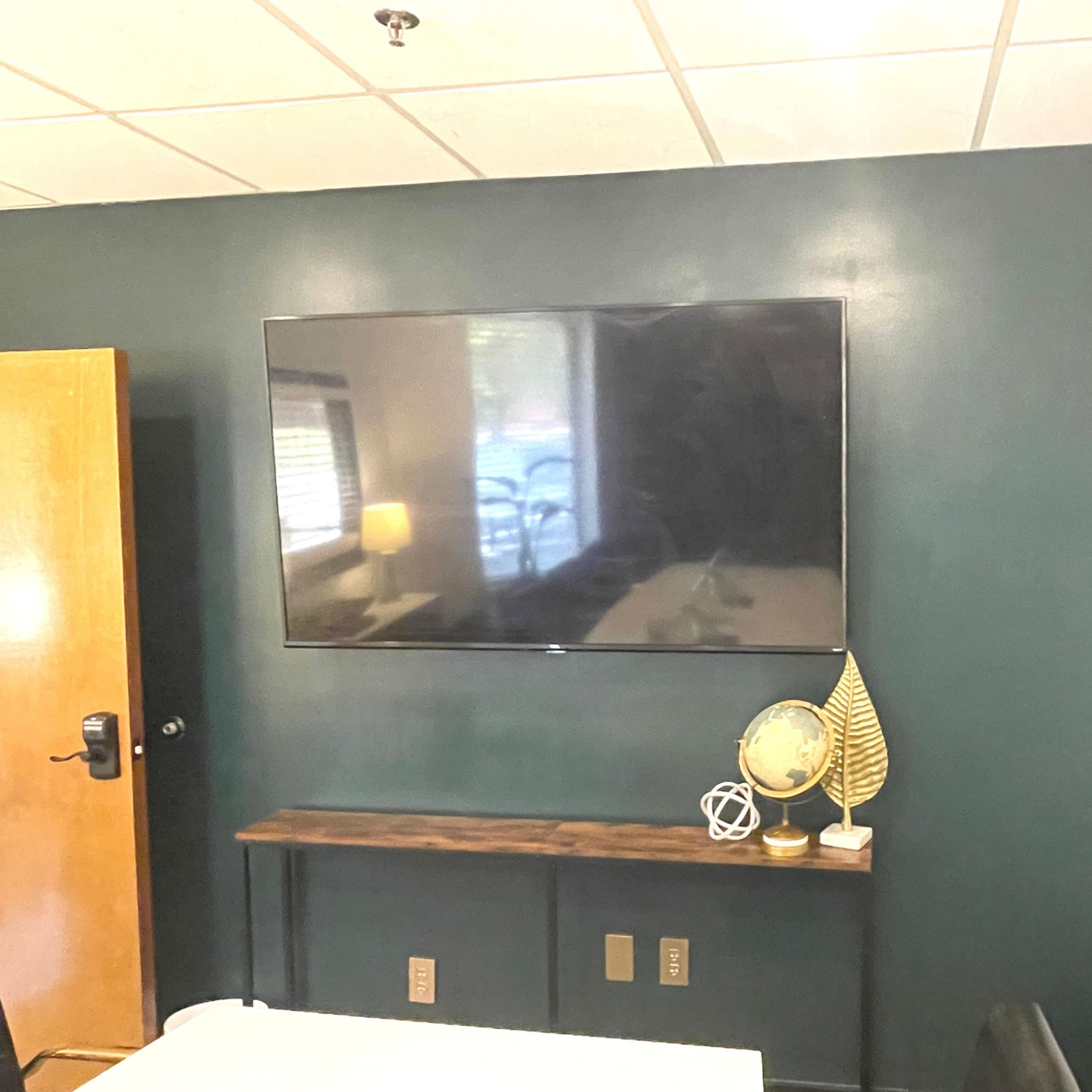 A large television is mounted on a dark green wall above a wooden console table, with a decorative globe and a leaf sculpture on top.
