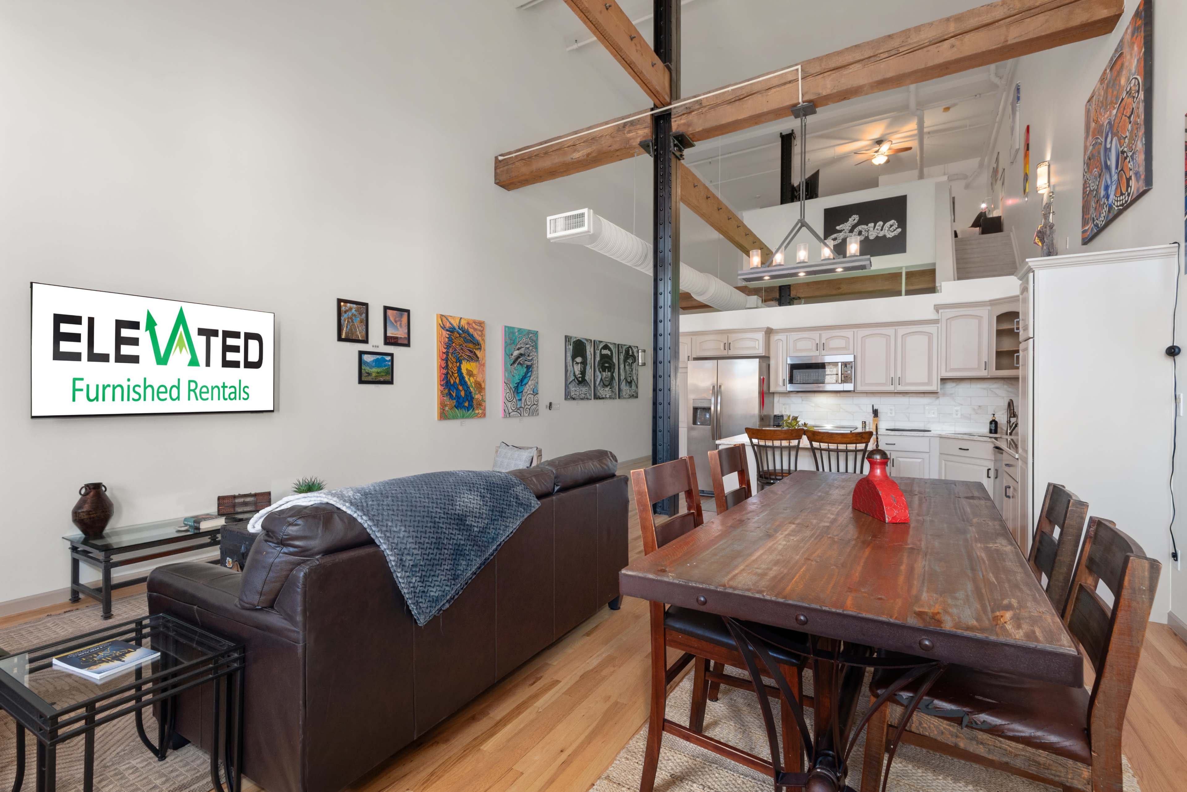 A spacious living area features a leather sofa, a wooden dining table, and a wall displaying various artworks, with a sign for "Elevated Furnished Rentals" visible.