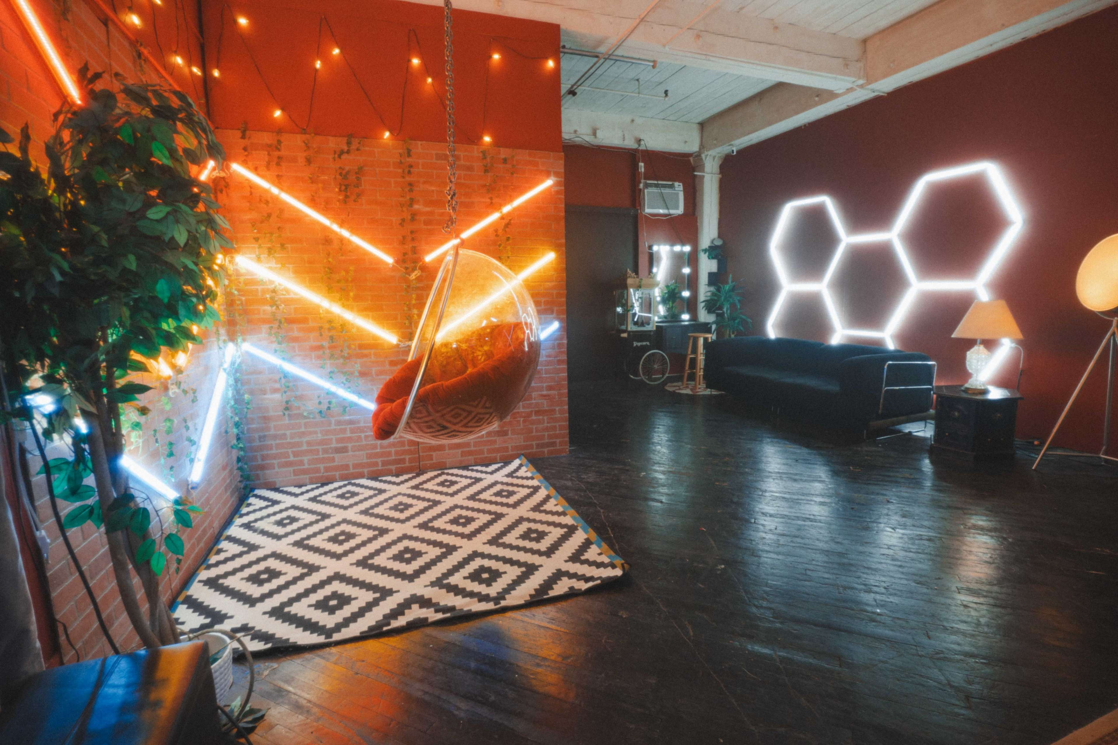 The image shows a modern interior space featuring a hanging chair, geometric lighting, a patterned rug, and minimal furniture against a brick wall.