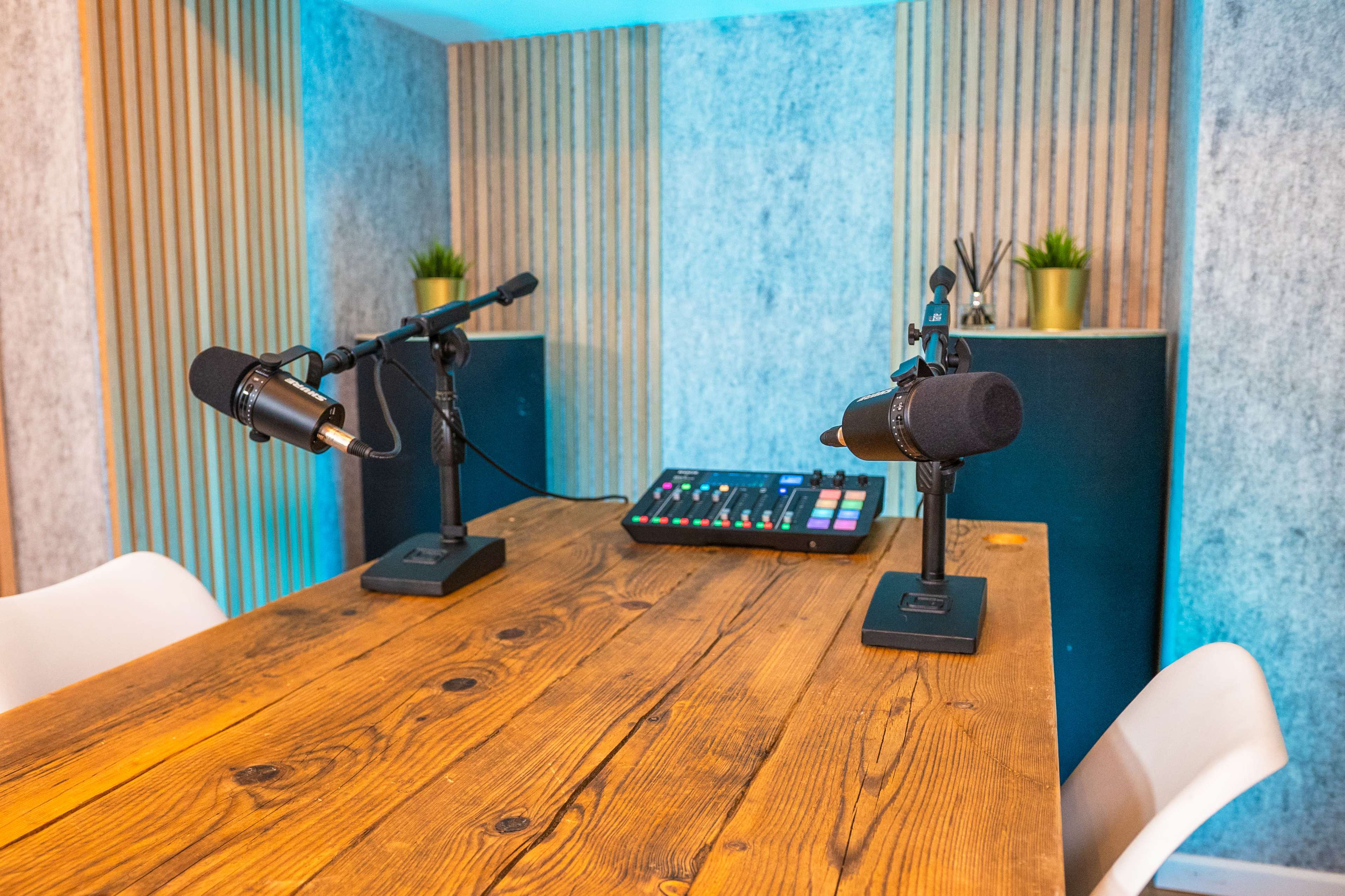 A small podcast studio with two microphones on stands, a wooden table, and a soundboard with colorful buttons set against a backdrop of textured walls and small plants.