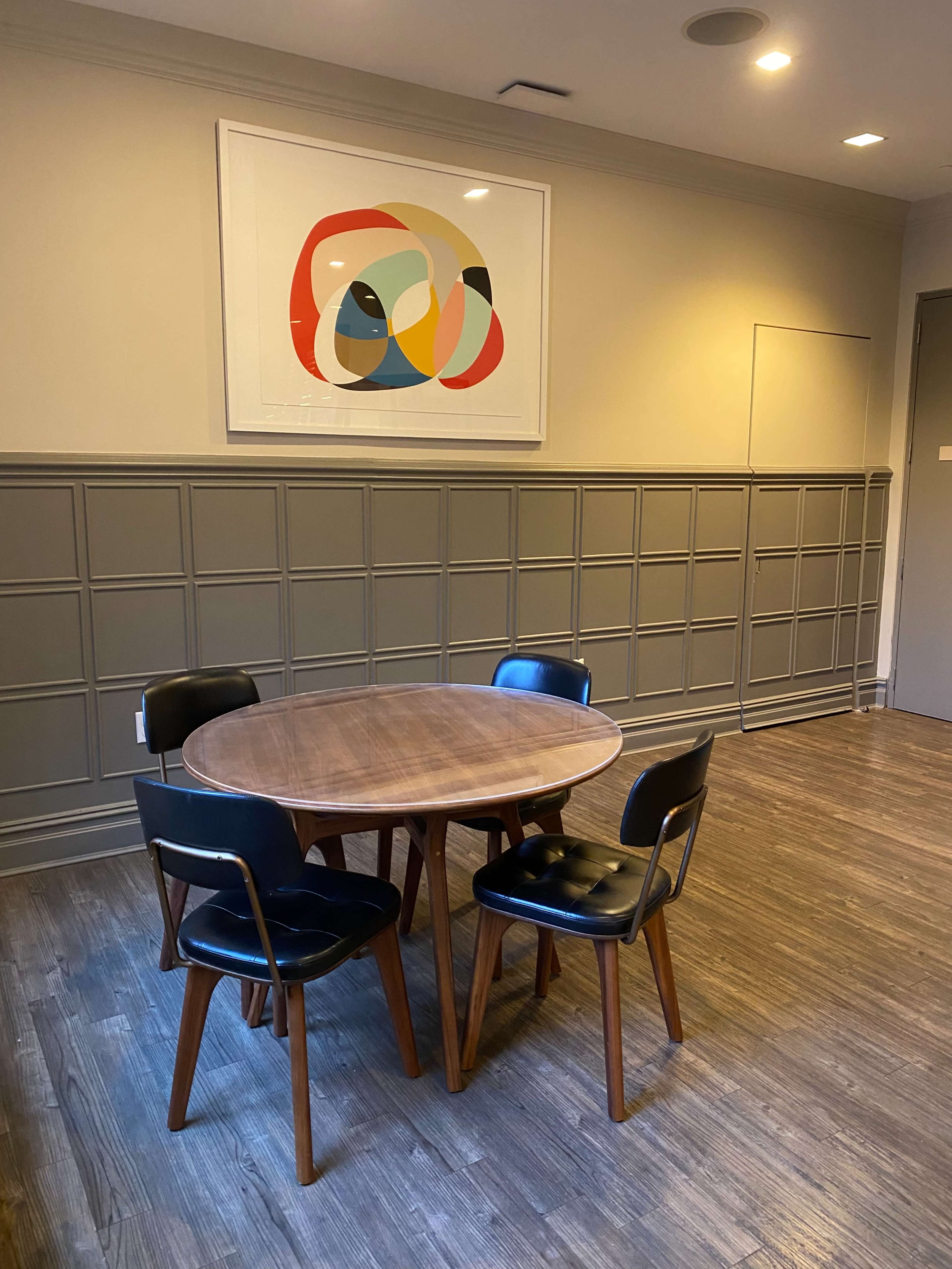 A small dining area with a round wooden table surrounded by four black chairs, situated against a paneled wall and featuring a colorful abstract artwork above.