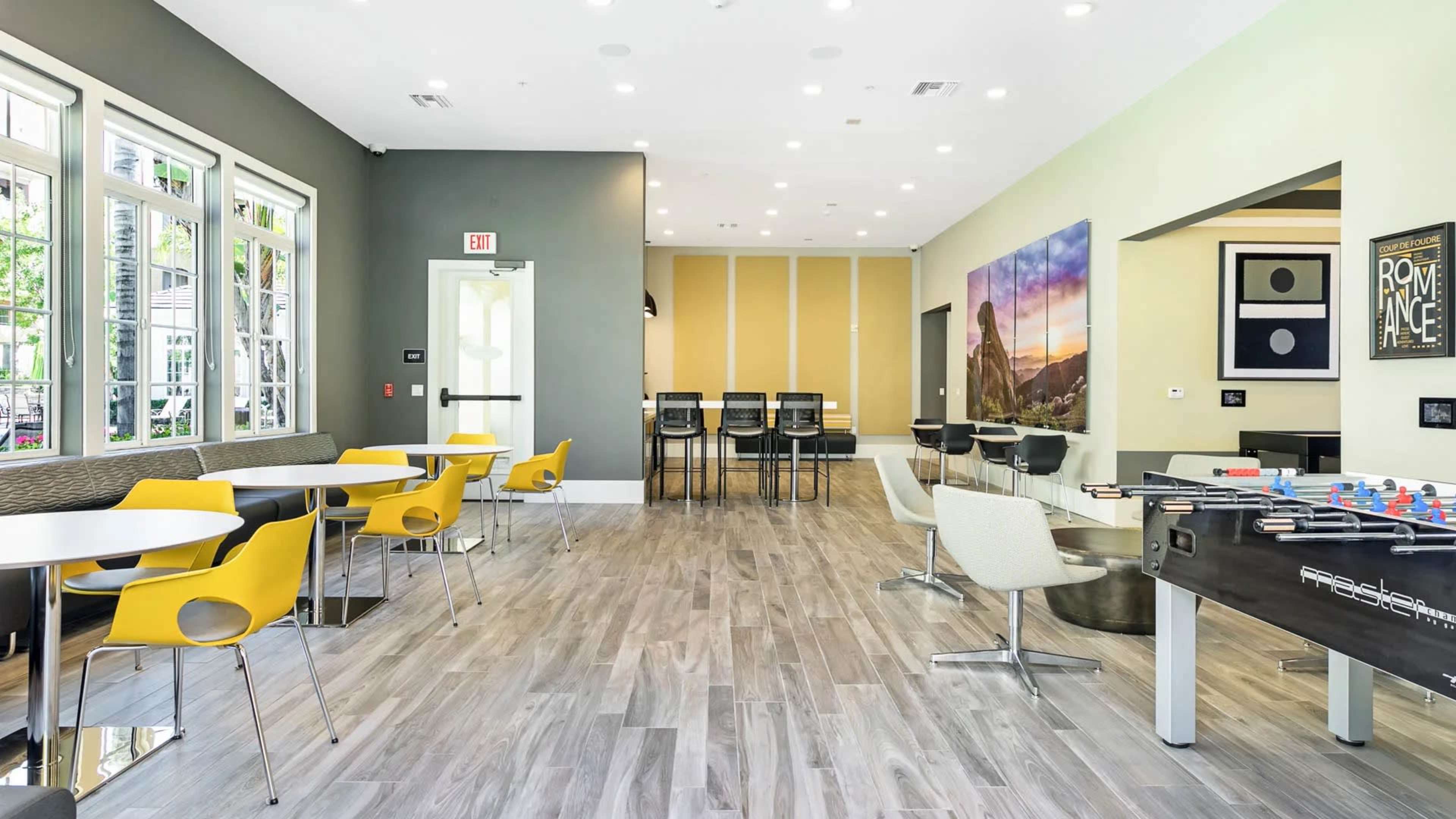 A modern common area with tables, chairs, and a foosball table, featuring large windows and light-colored walls.