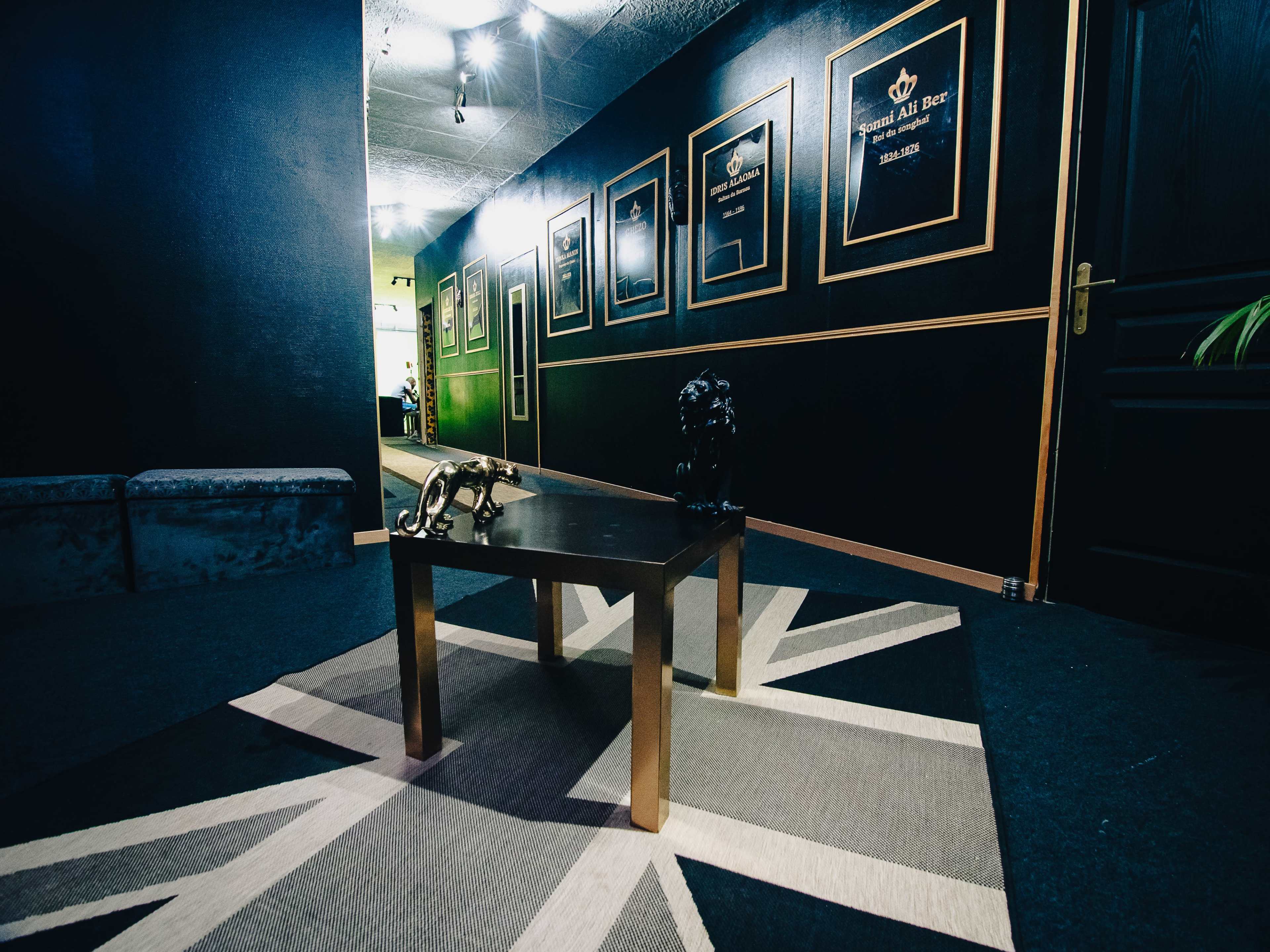 A hallway with black walls, a patterned rug featuring a Union Jack design, a small table with decorative figures, and framed artwork or plaques on the walls.