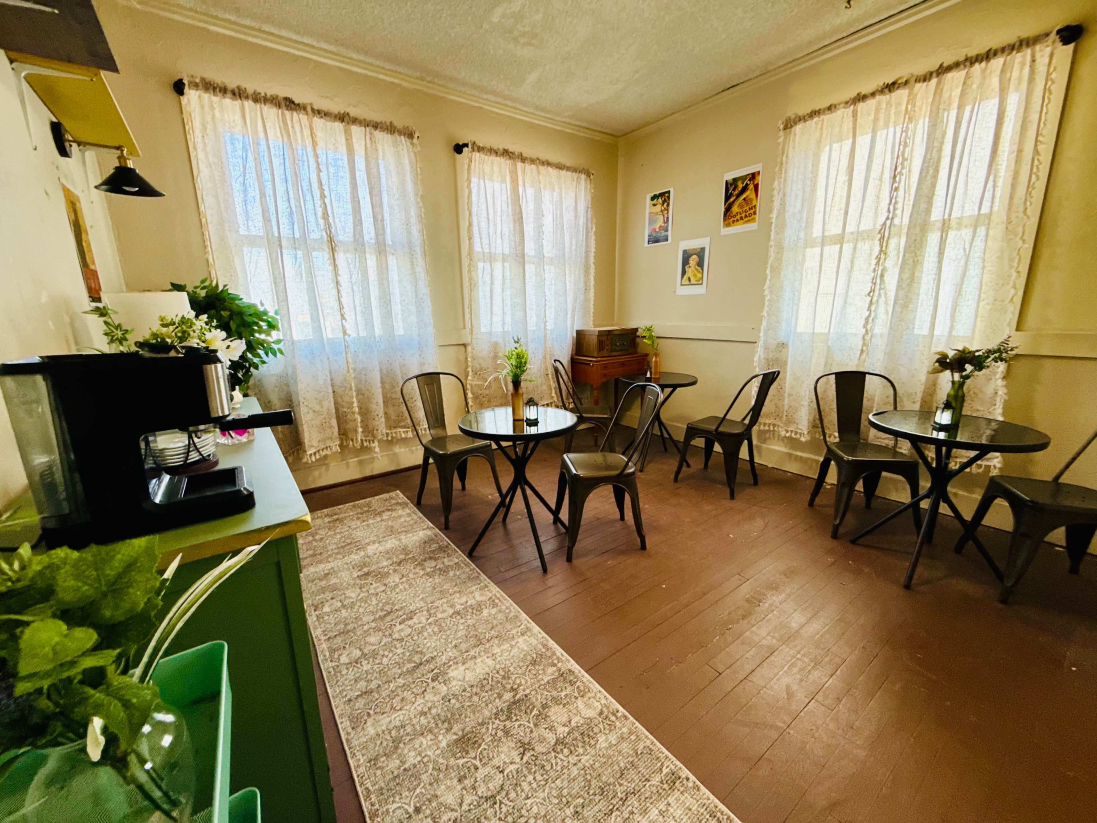 The image shows a cozy room with two tables, black chairs, large windows adorned with lace curtains, and a decorative side table.
