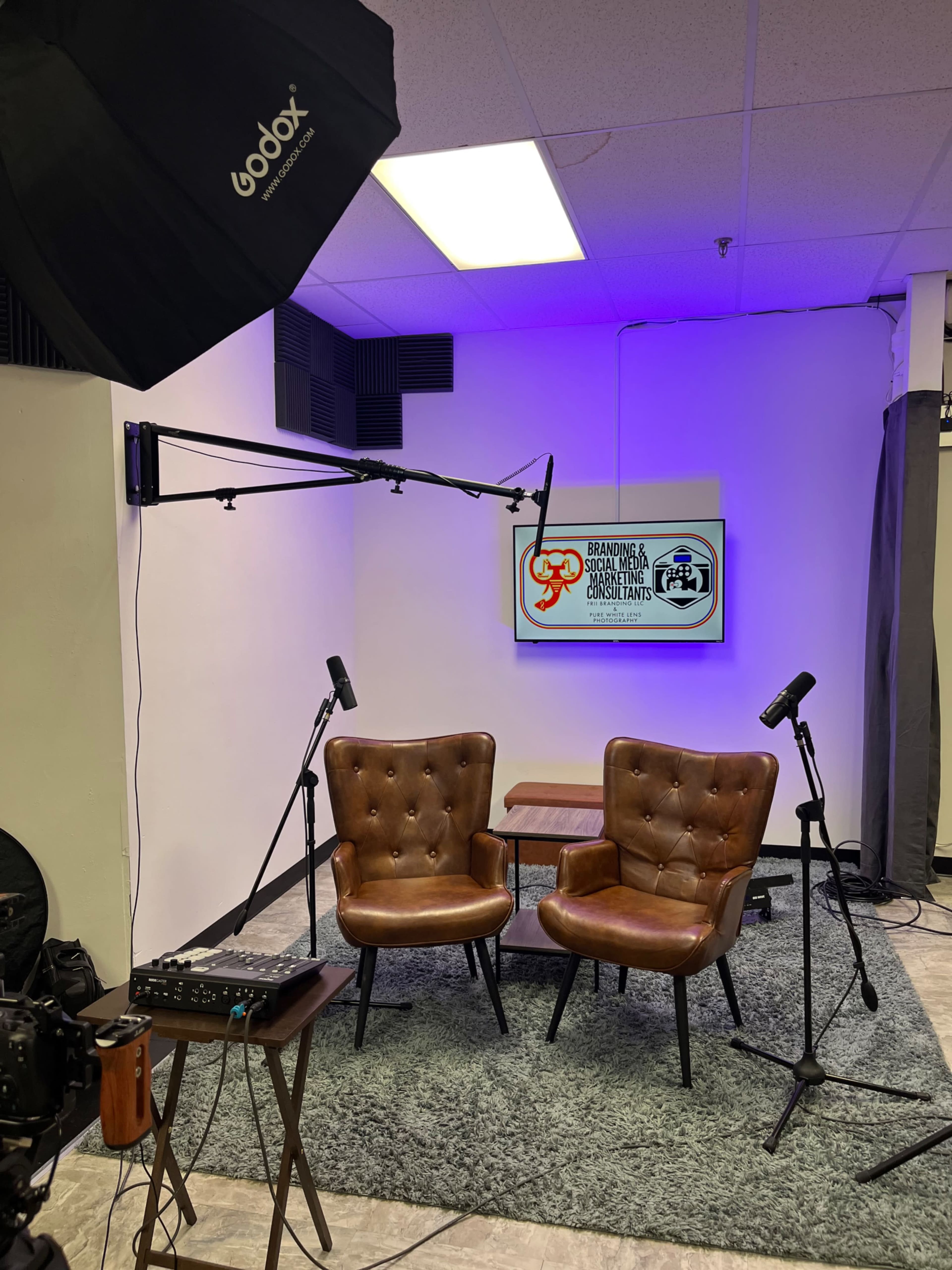 A small studio setup with two brown chairs, a table between them, and a screen displaying logos in the background, along with video equipment and lighting.