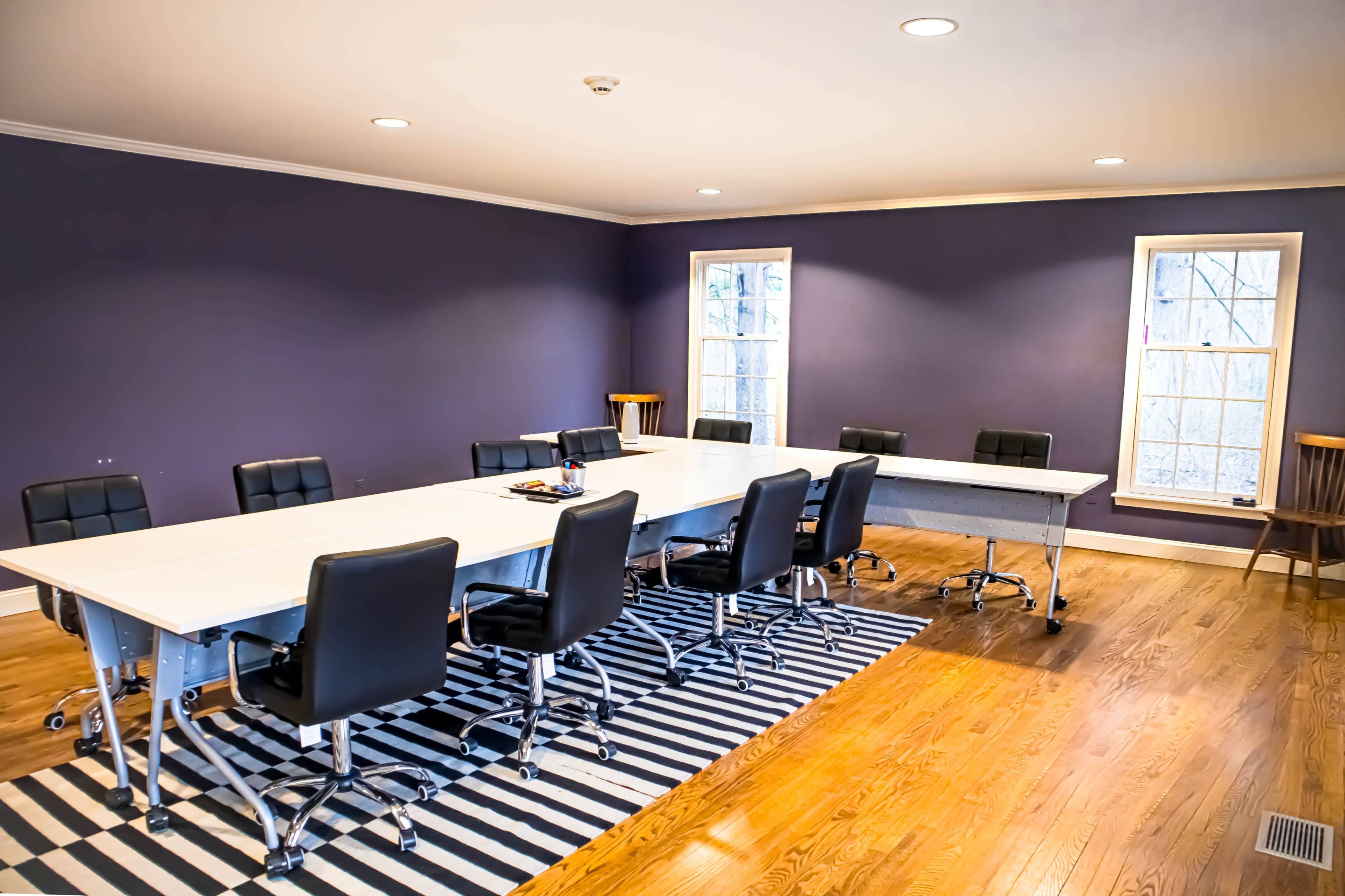 A spacious conference room features a long white table surrounded by black chairs on a striped rug with windows letting in natural light.