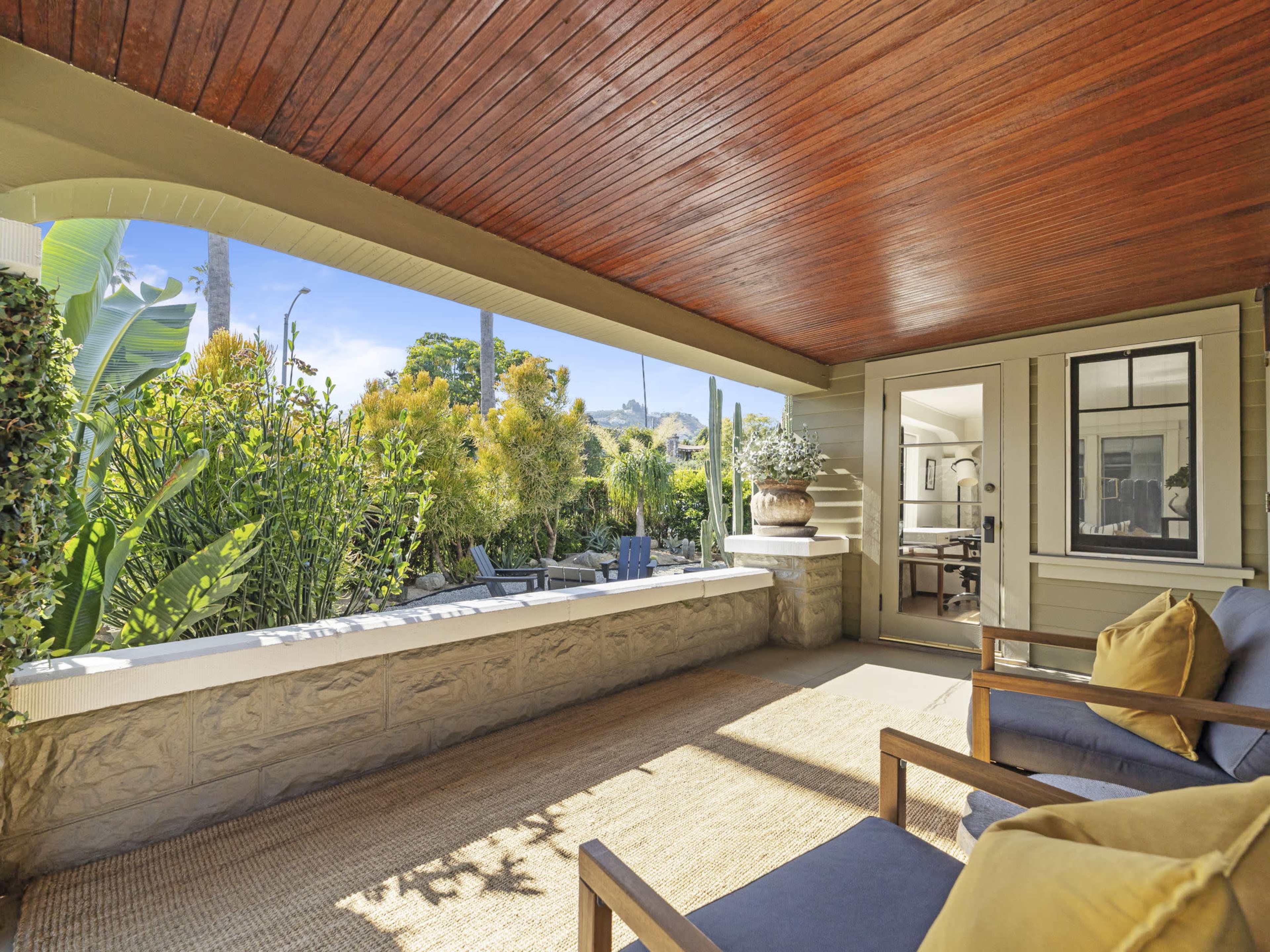 The image shows a covered porch with wooden seating, a textured rug, and a view of a lush garden beyond.