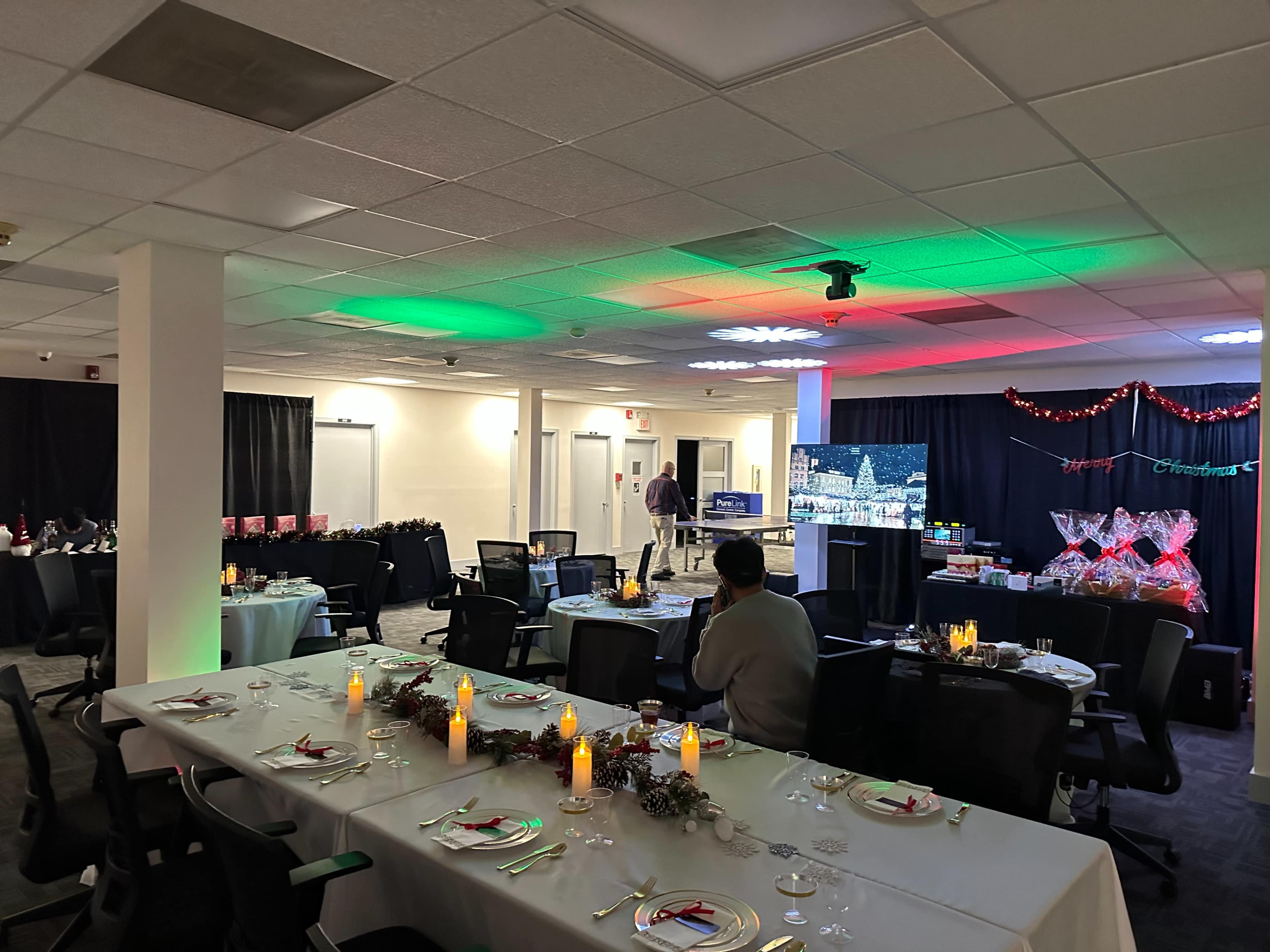 A spacious room is set up for a festive gathering, featuring decorated tables with candles and a backdrop of holiday-themed decorations.