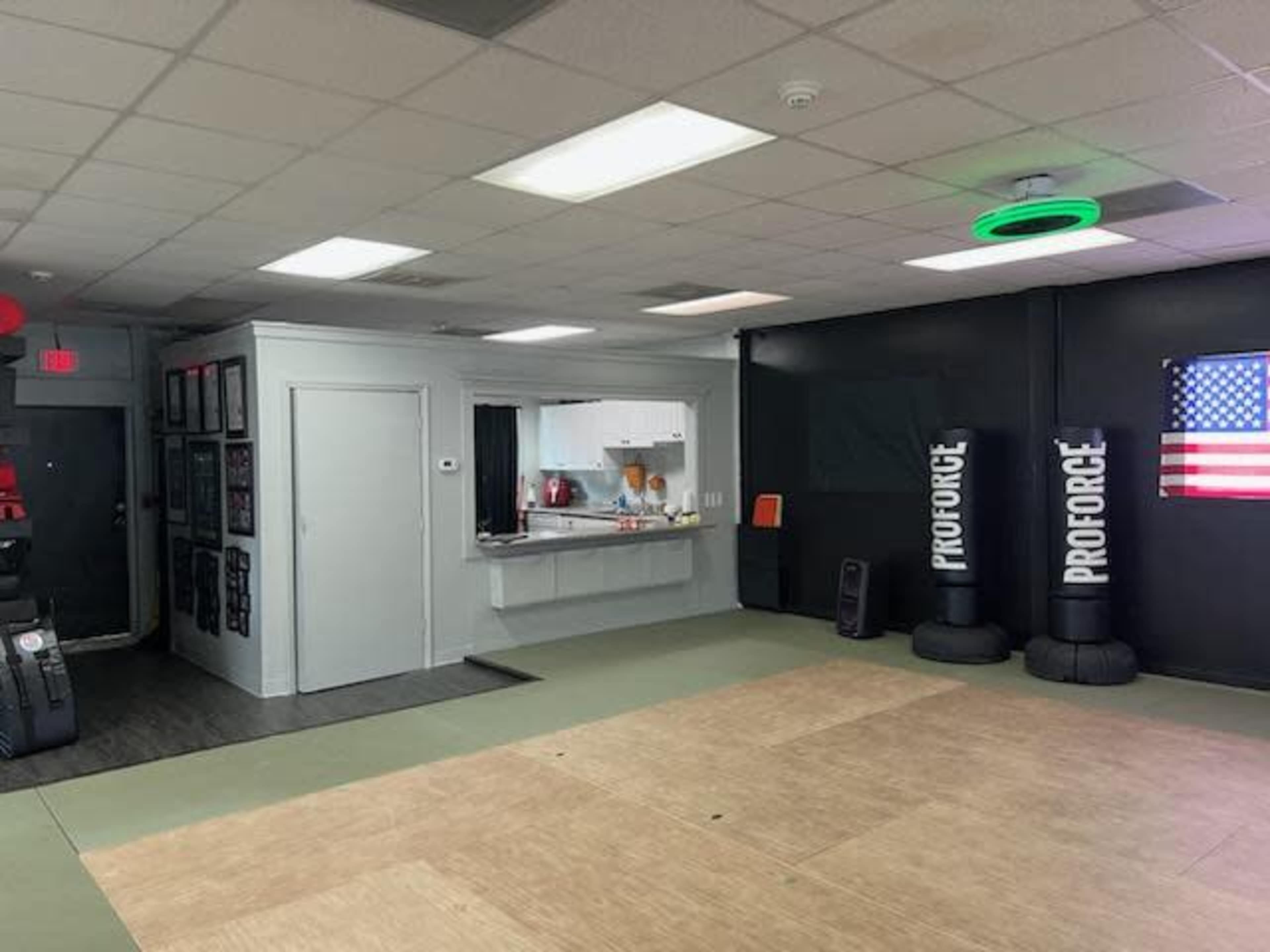 The image shows an interior gym space with exercise mats, a kitchen area visible through an opening, and two punching bags against a dark wall featuring an American flag.