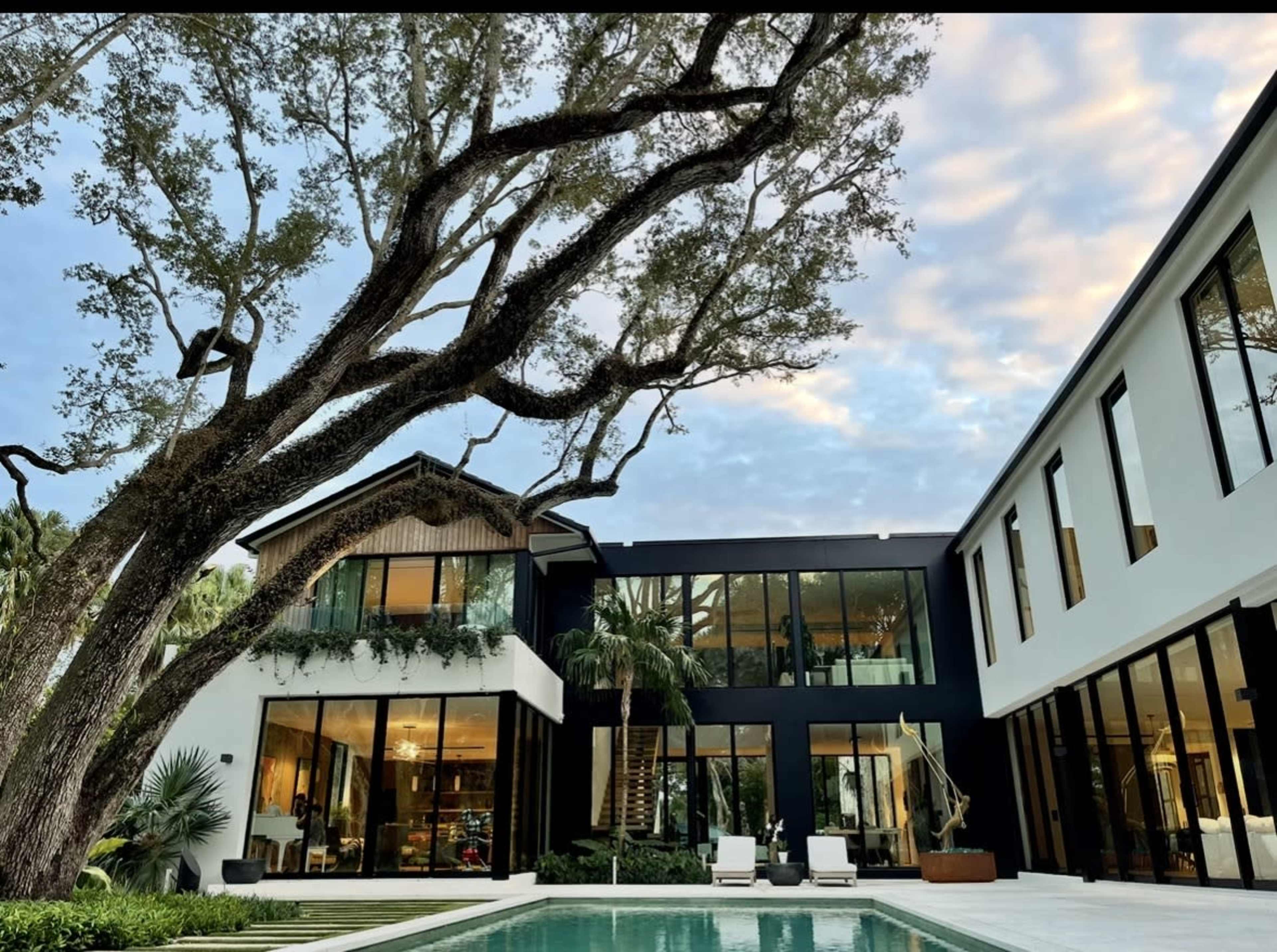 The image depicts a modern house with large glass windows, surrounded by greenery and a swimming pool in the foreground.