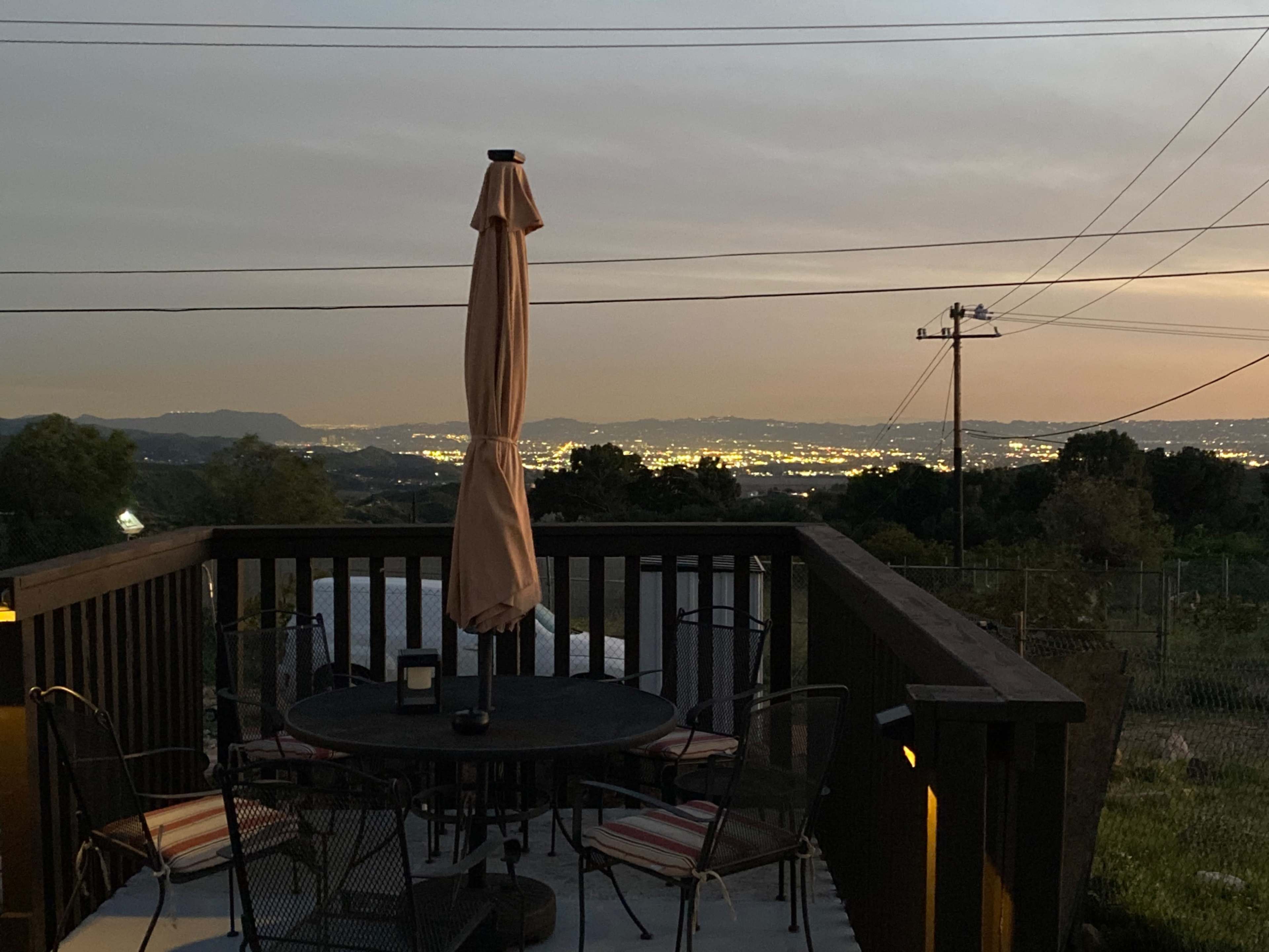 A patio with a table and chairs overlooks a city lit by evening lights against a scenic backdrop of mountains.