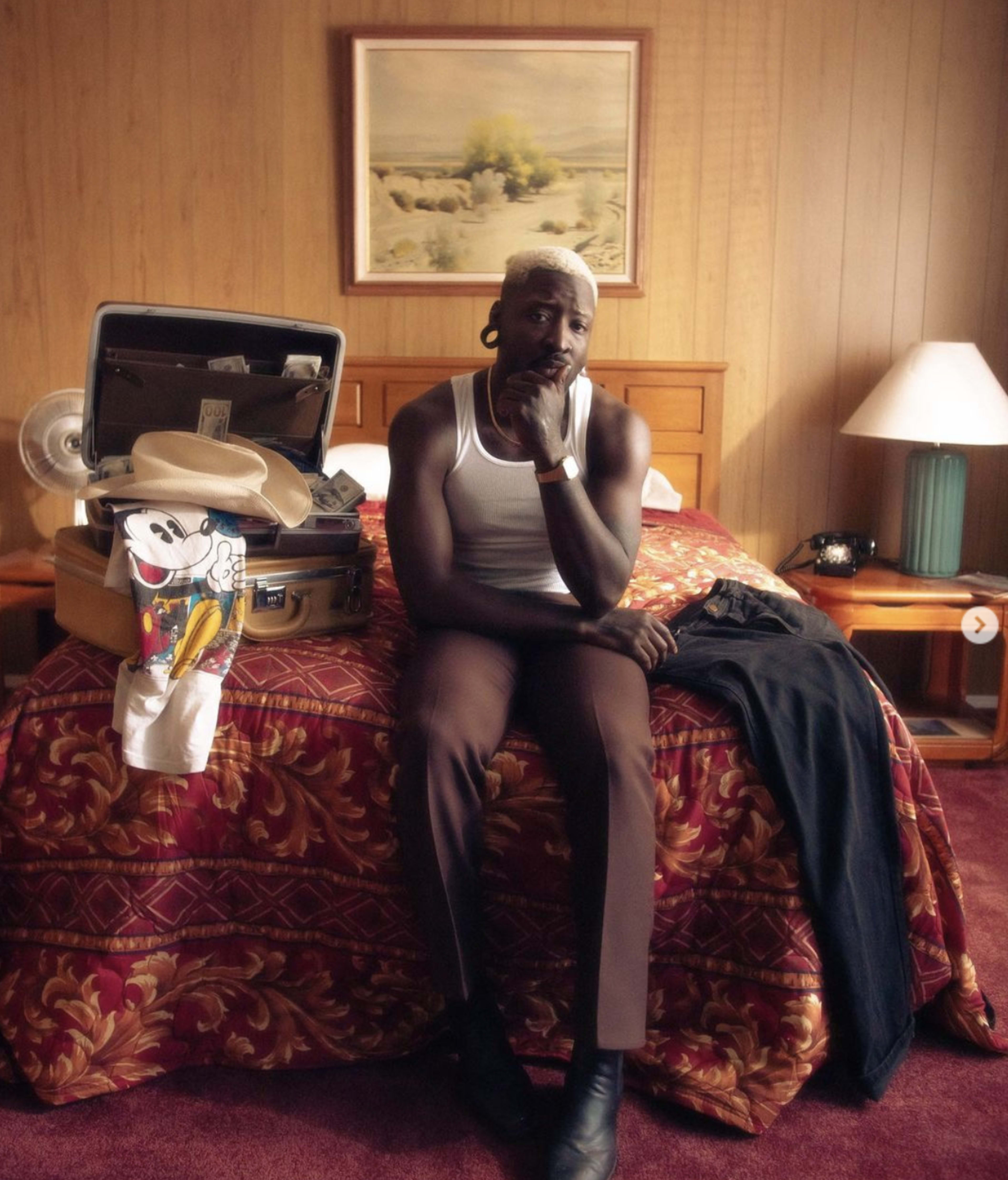 A person sits on a bed with a suitcase open beside them, surrounded by clothing and a lamp in a motel room.