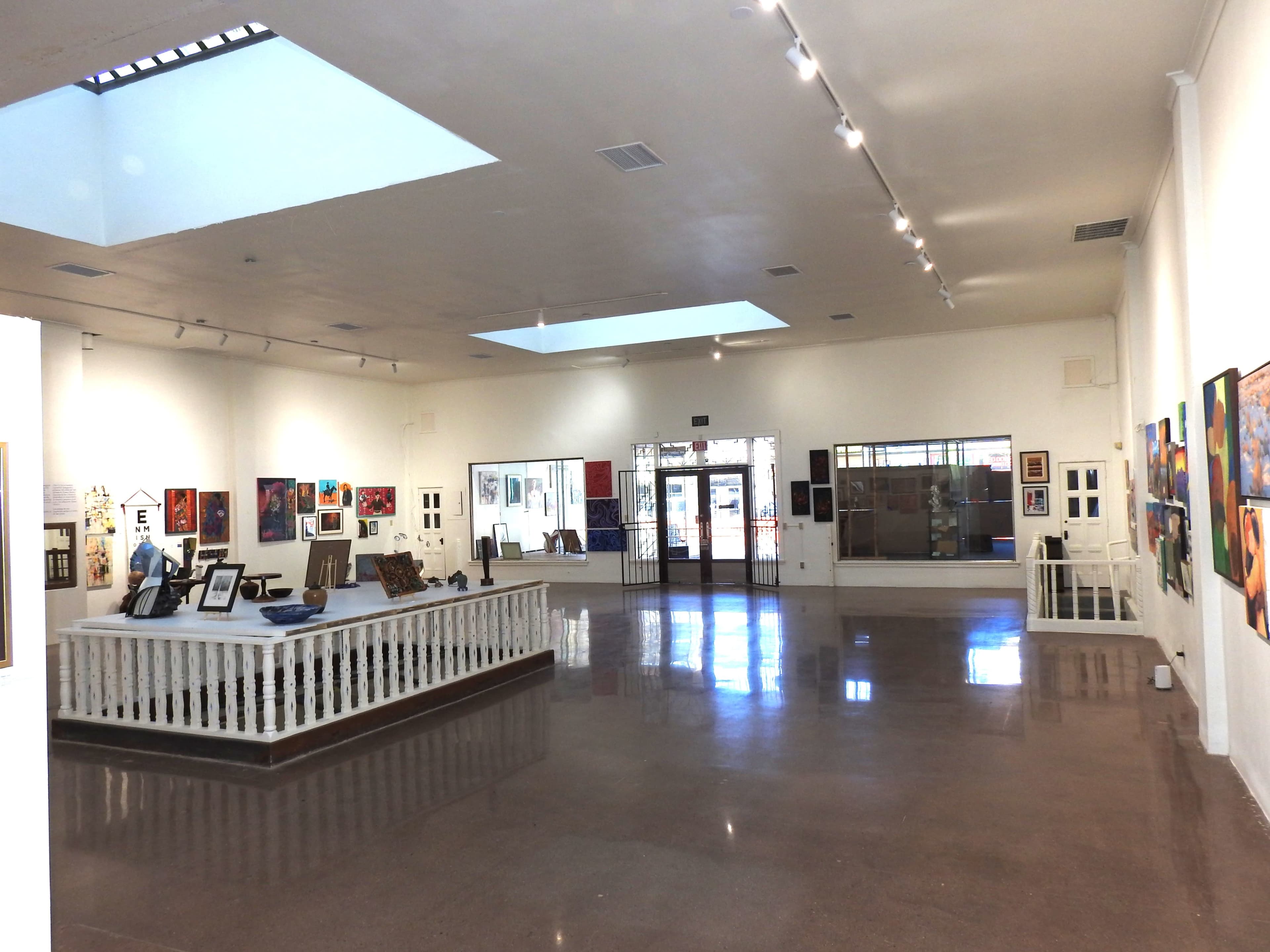 The image shows the interior of an art gallery featuring various paintings on the walls and a spacious layout with natural light coming from skylights.
