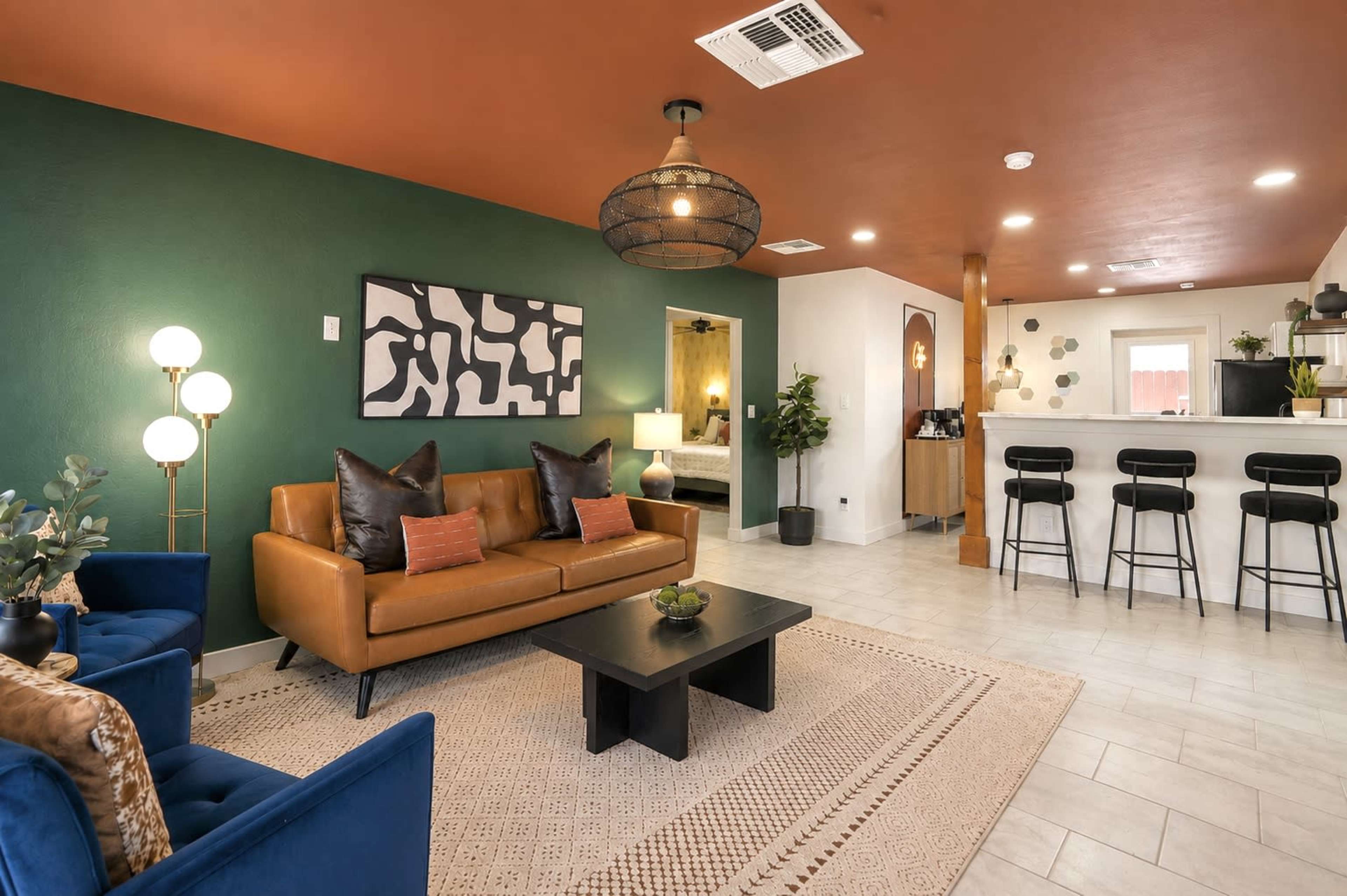 The image shows a modern living area with a brown leather sofa, blue accent chairs, a black coffee table, and a bar area with high stools, all set against a backdrop of green and orange walls.
