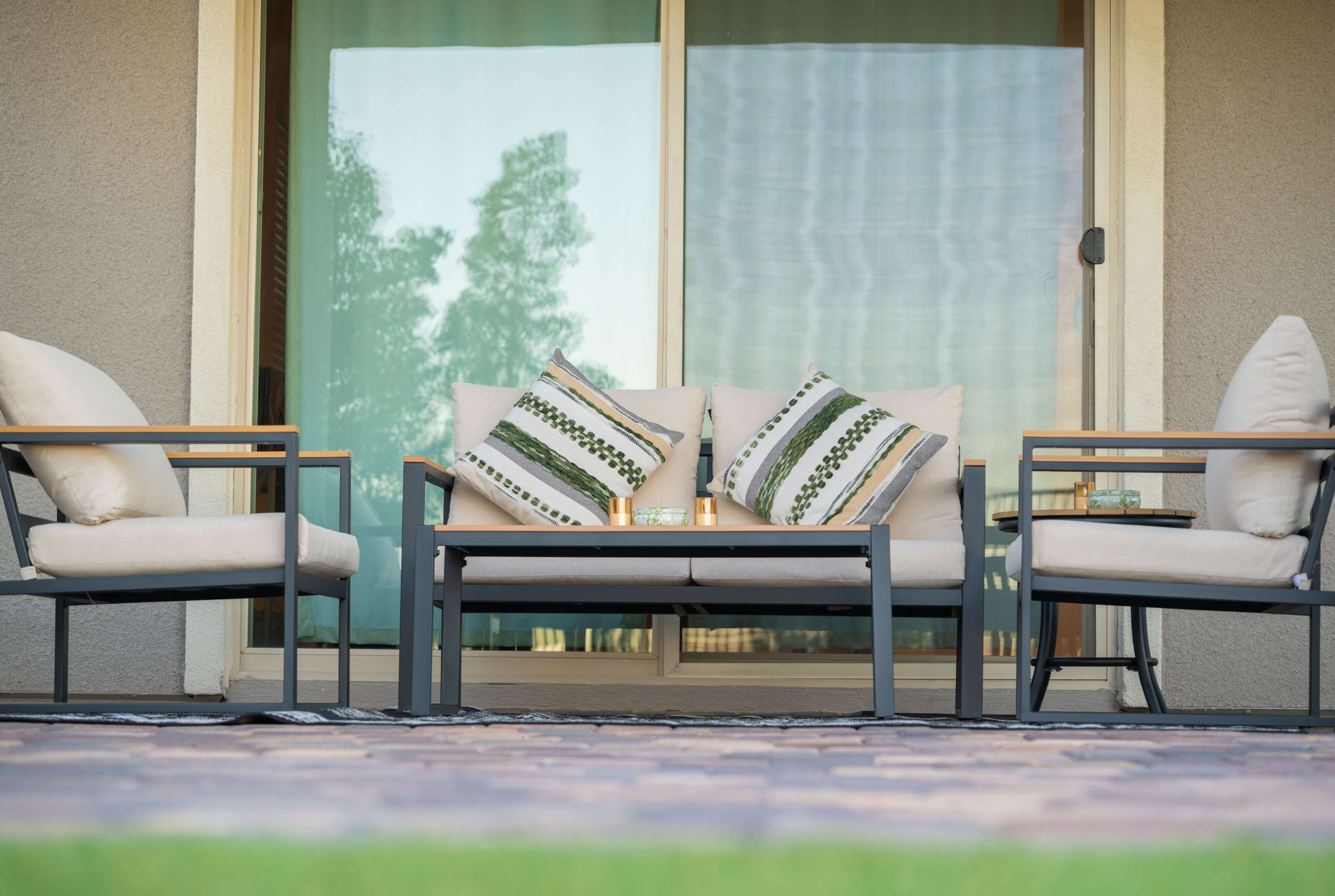 A set of outdoor furniture, including a sofa with decorative pillows and two armchairs, is arranged on a patio in front of a large glass window.