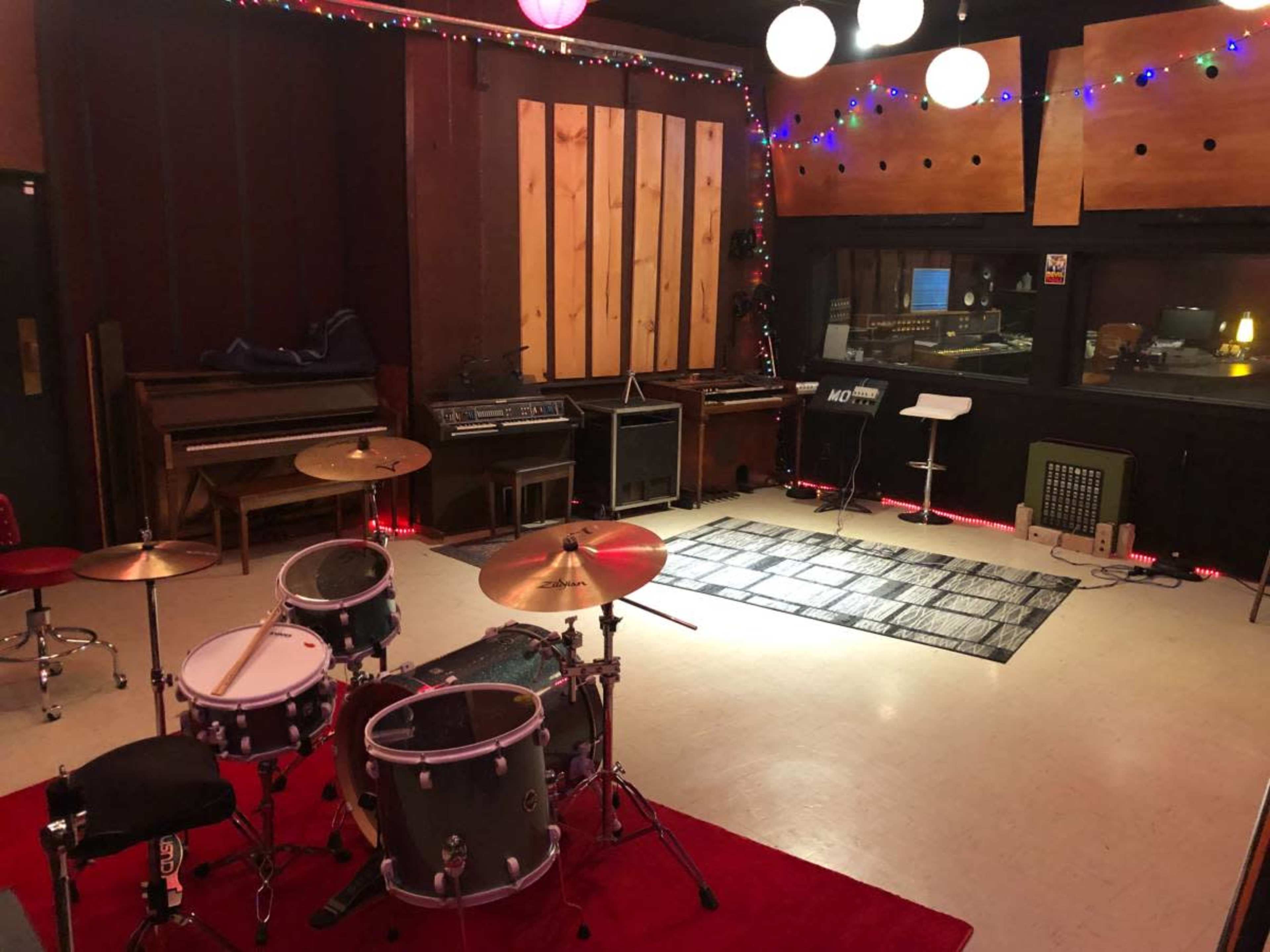 The image shows a music studio with a drum set, keyboard, amplifiers, and a control room visible behind glass.