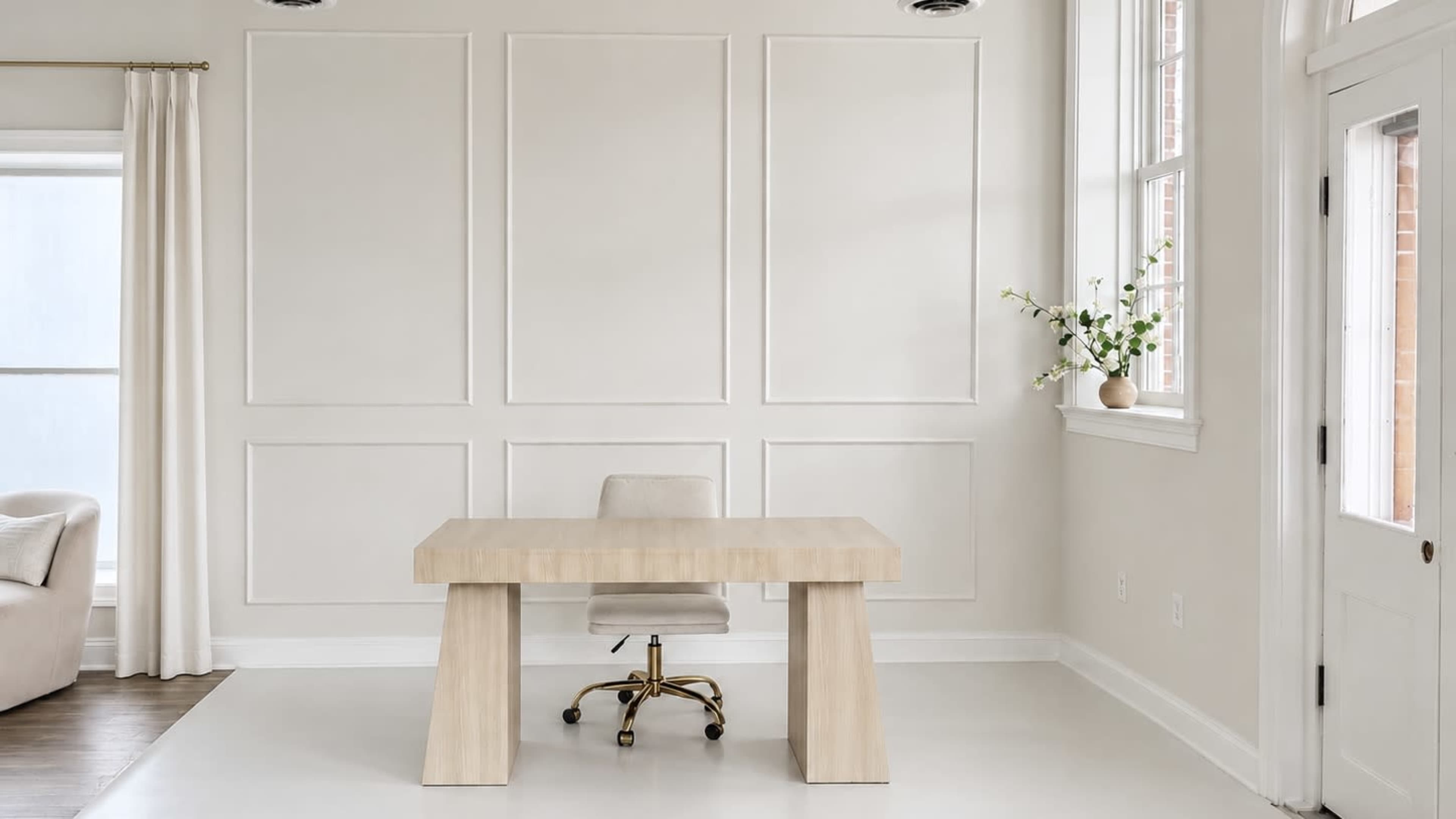 The image shows a minimalist office space featuring a wooden desk with a rolling chair against a light-colored wall with paneling.