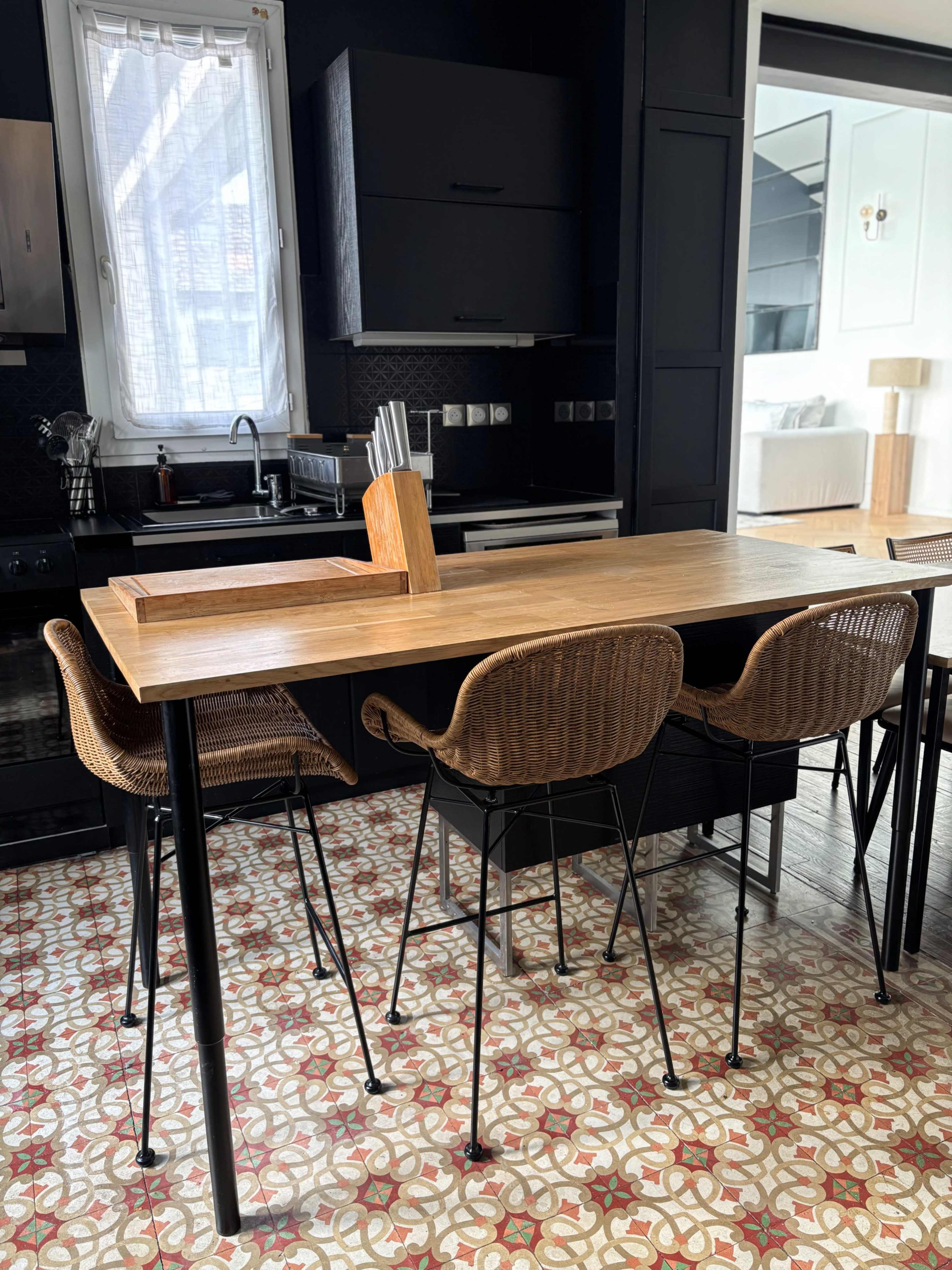The image shows a modern kitchen with a wooden table, black cabinetry, and woven chairs, featuring patterned tiles on the floor.