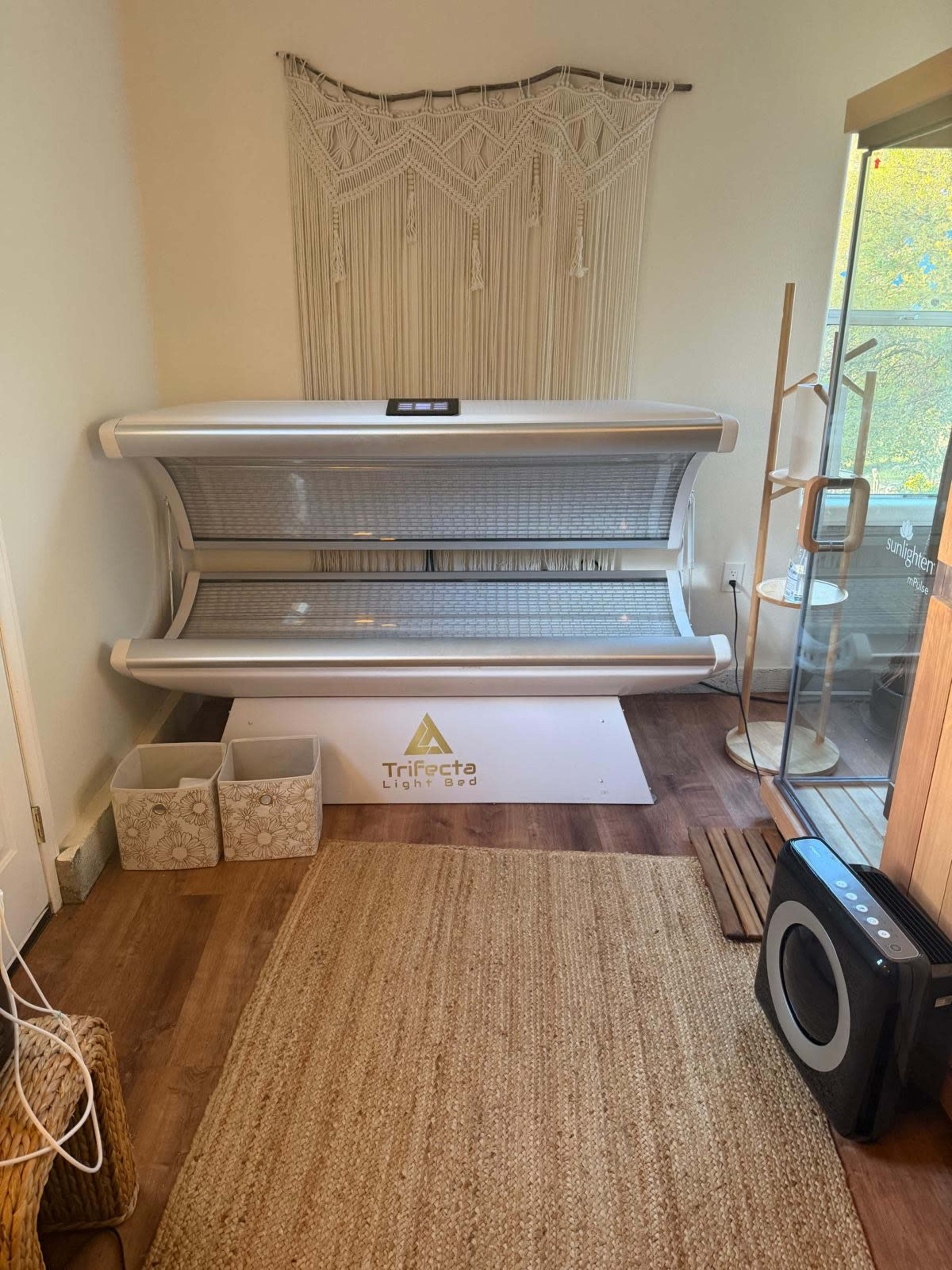 The image shows a tanning bed positioned in a well-lit room decorated with a wall hanging and a natural fiber rug.