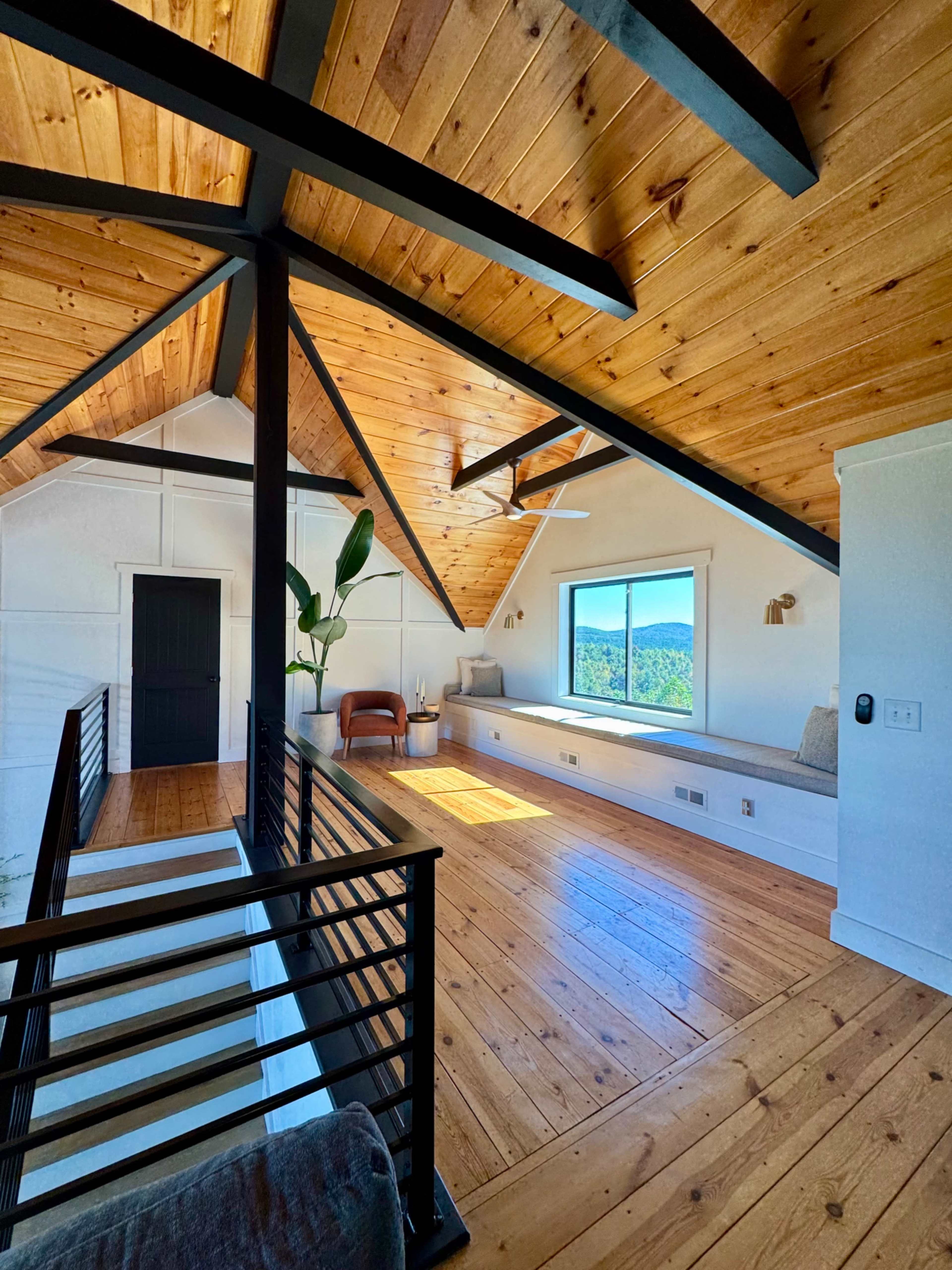 The image shows a bright, open loft space with hardwood floors, wooden beams, and a large window offering mountain views.