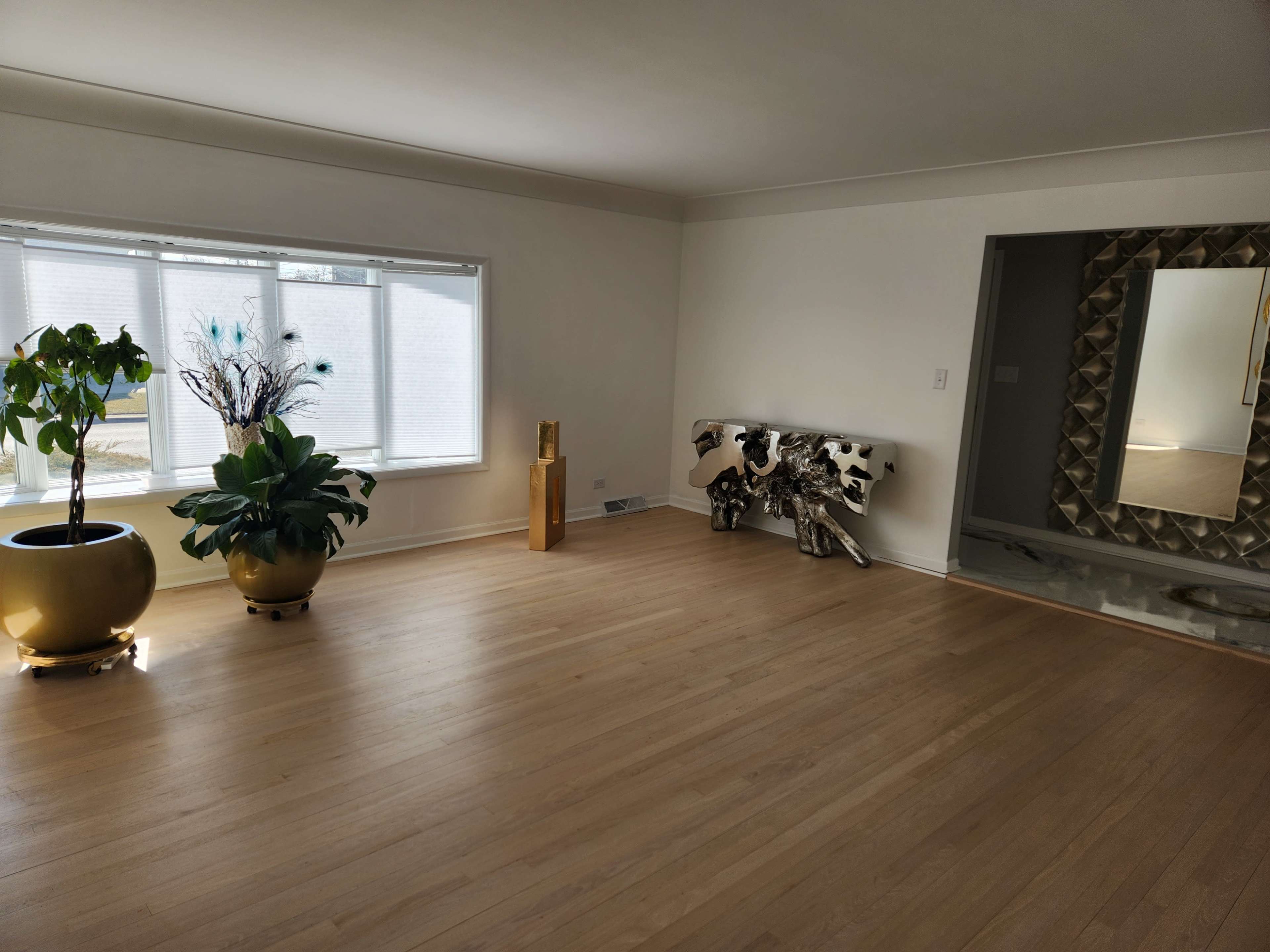 The image shows a spacious, minimally furnished living room with large windows, wooden floors, and decorative plants.