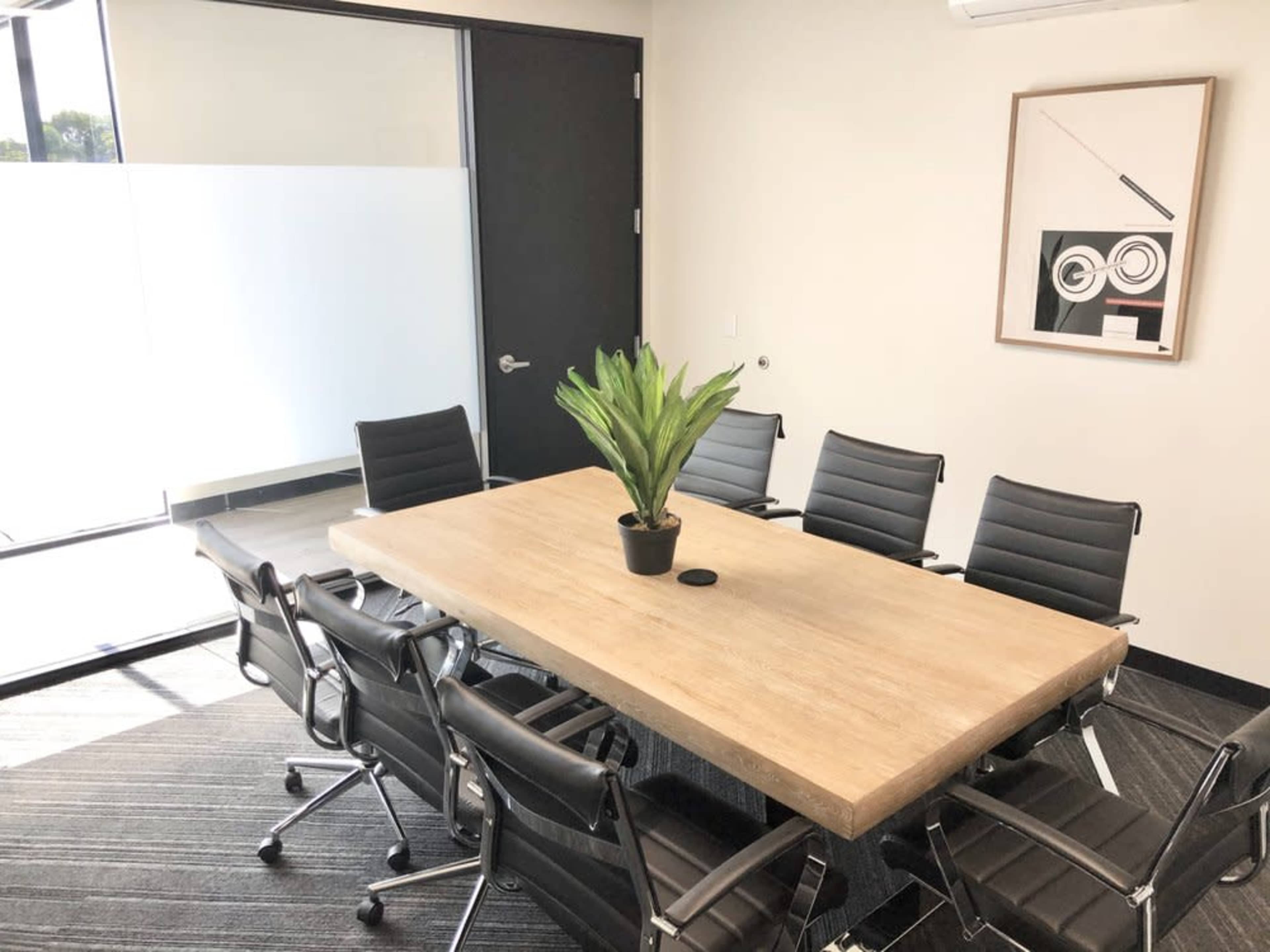 The image shows a modern meeting room with a large wooden table surrounded by black rolling chairs and a green plant centerpiece.