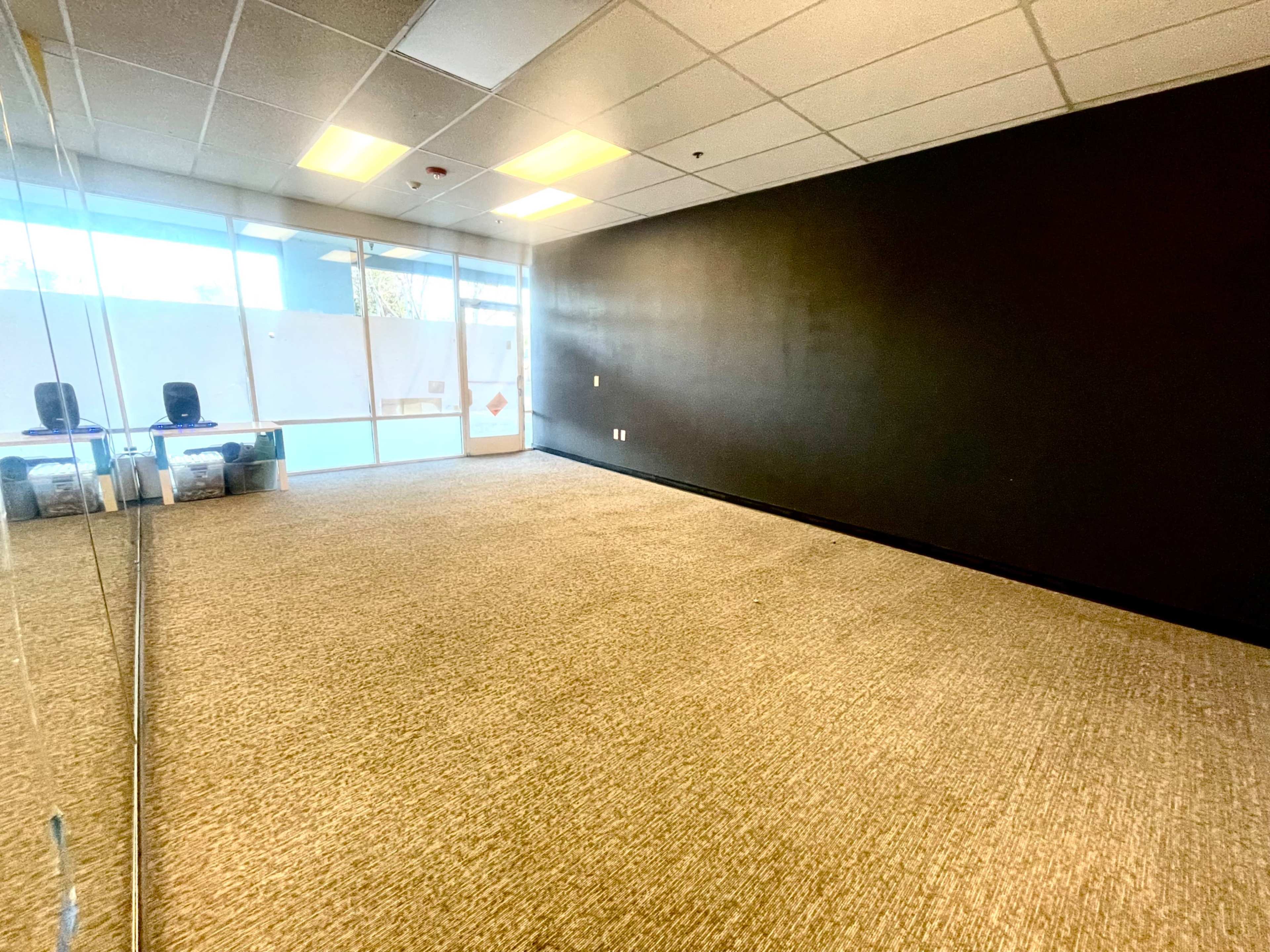A spacious, empty room with a dark wall and a large mirror on one side, featuring a carpeted floor and window light coming from the entrance.
