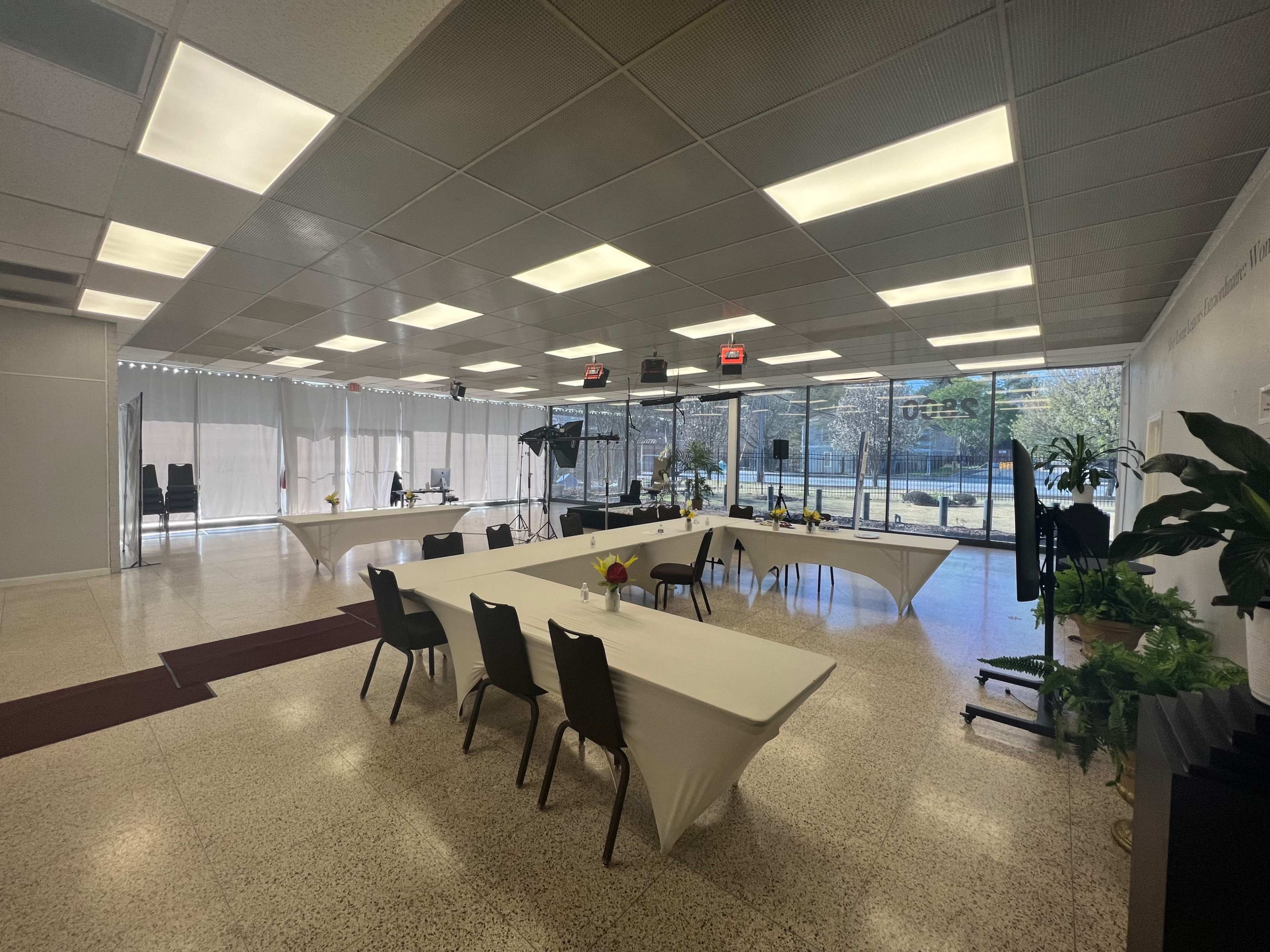 The interior of a spacious event room features multiple tables covered with white tablecloths and chairs arranged in a tidy layout, alongside large windows allowing natural light.