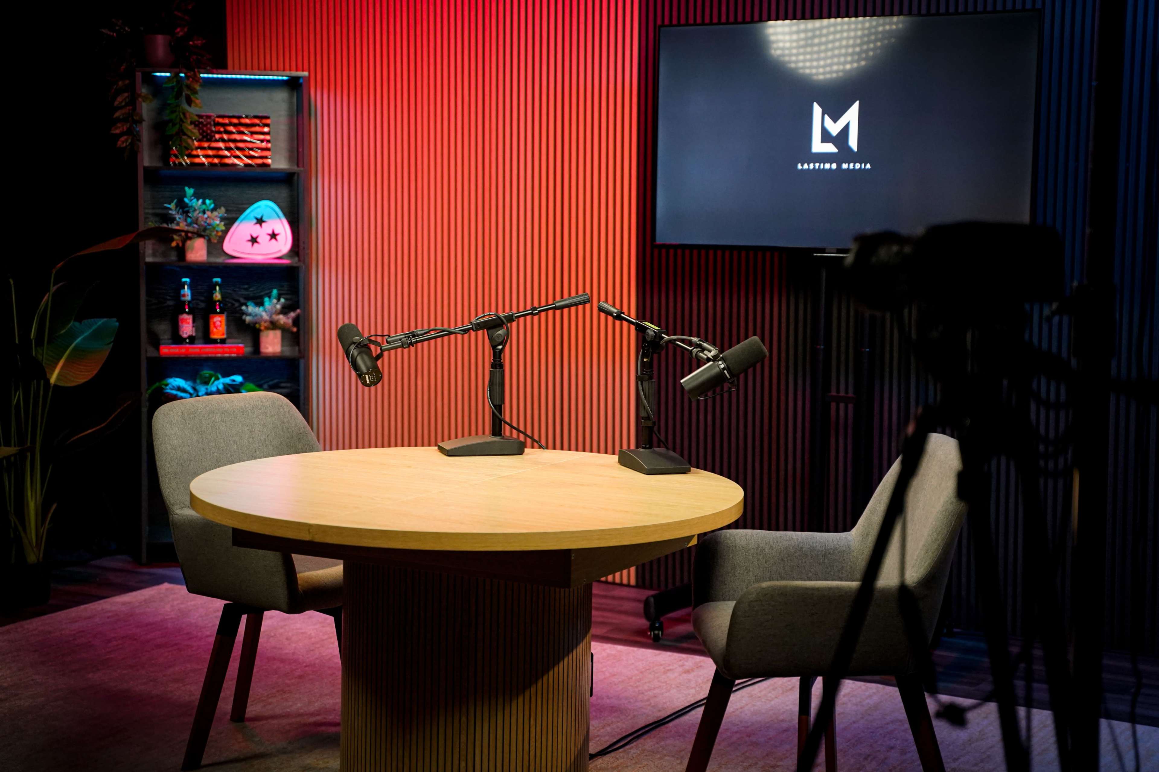 The scene shows a podcasting studio with two microphones positioned on a round table and a display screen featuring the logo "LATERAL MEDIA" in the background.