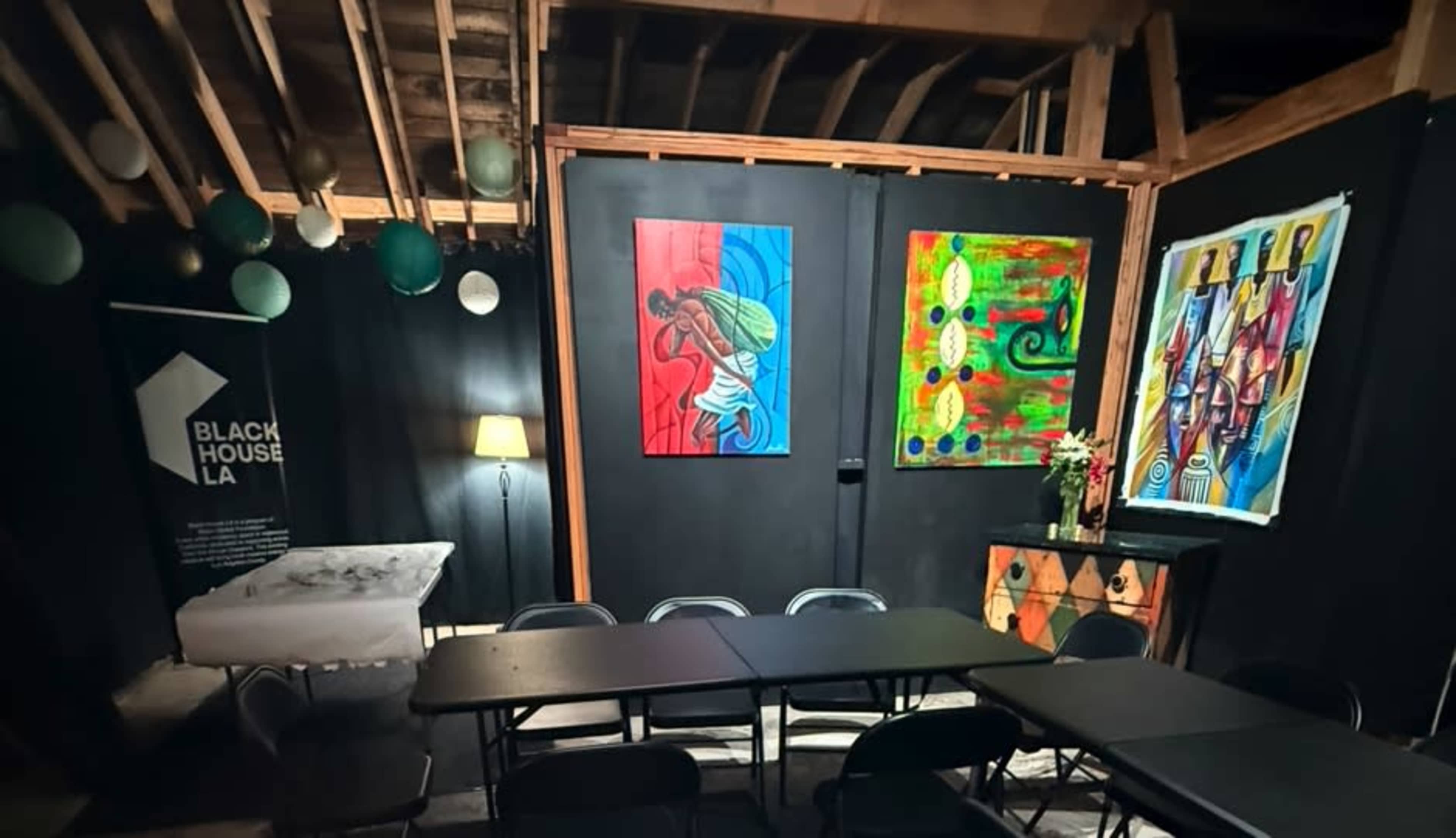 The image shows an indoor space with black walls, featuring two colorful artworks, a table with chairs, and a lamp in the corner.