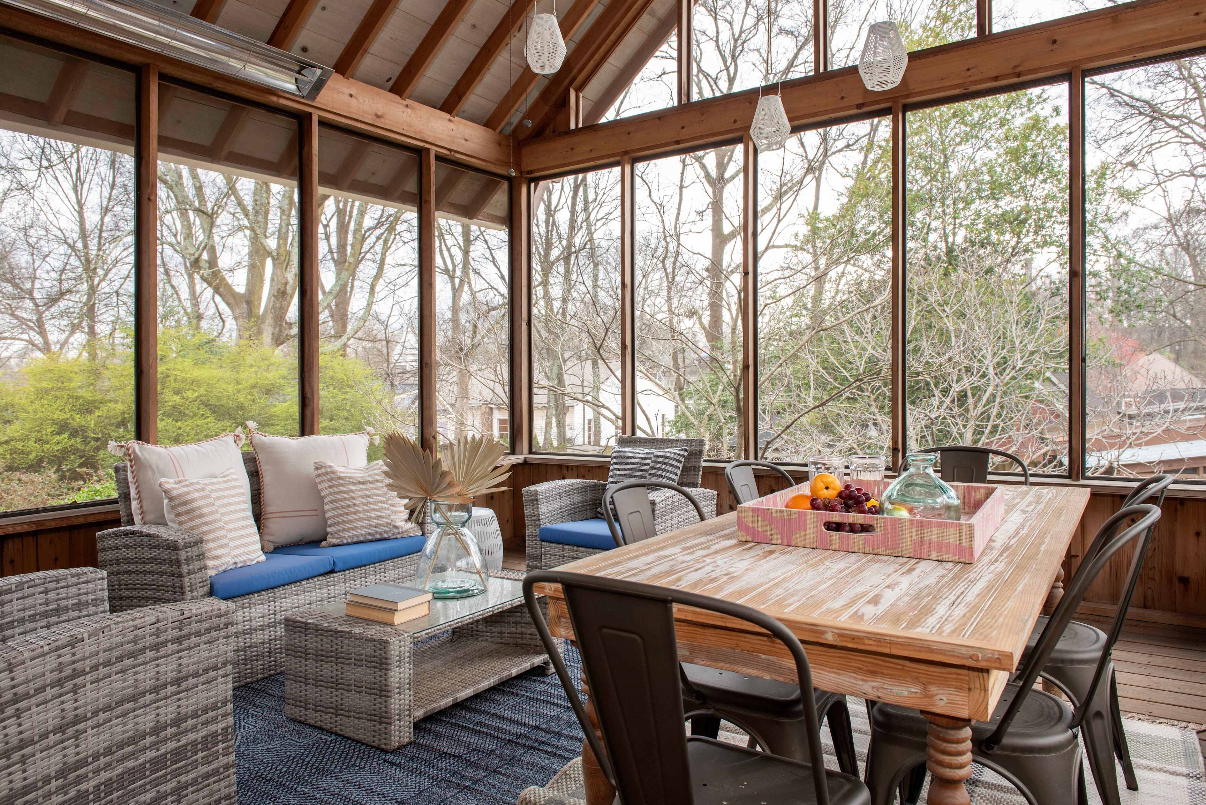The image shows a screened porch featuring a wooden dining table, wicker furniture, and large windows that provide views of a garden.