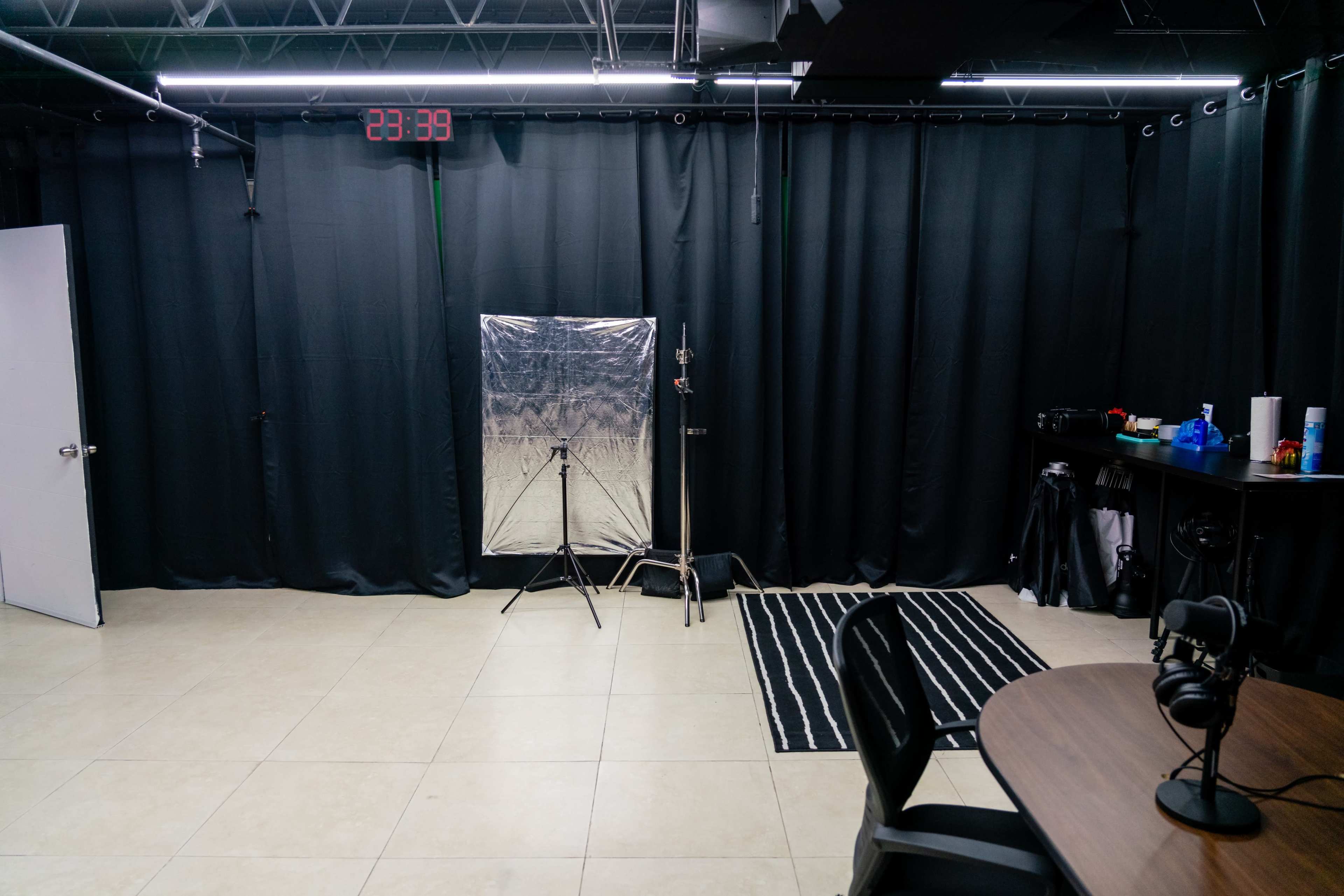 The image shows a photography studio with black curtains, a silver reflector on a stand, and a small table with microphones and various items along the wall.