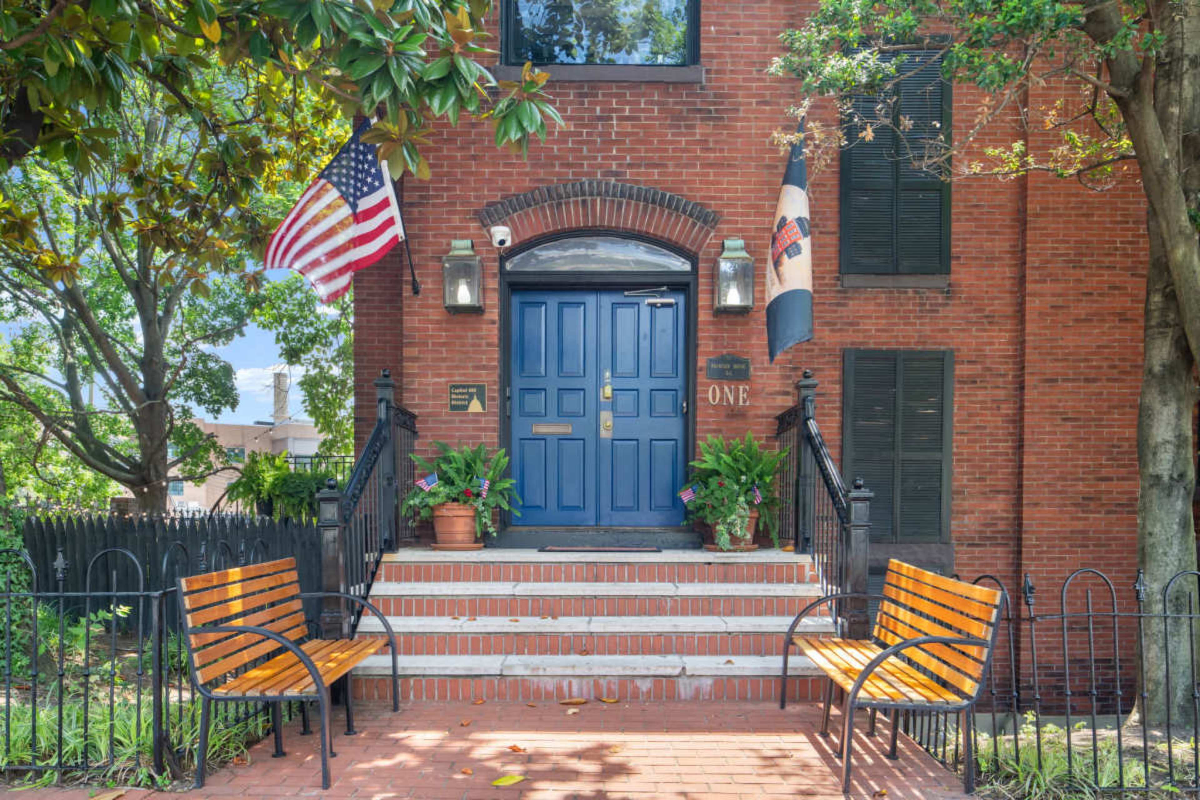 The image shows a brick front entrance with a blue door, flanked by American and state flags, and features two wooden benches on either side of a set of steps.