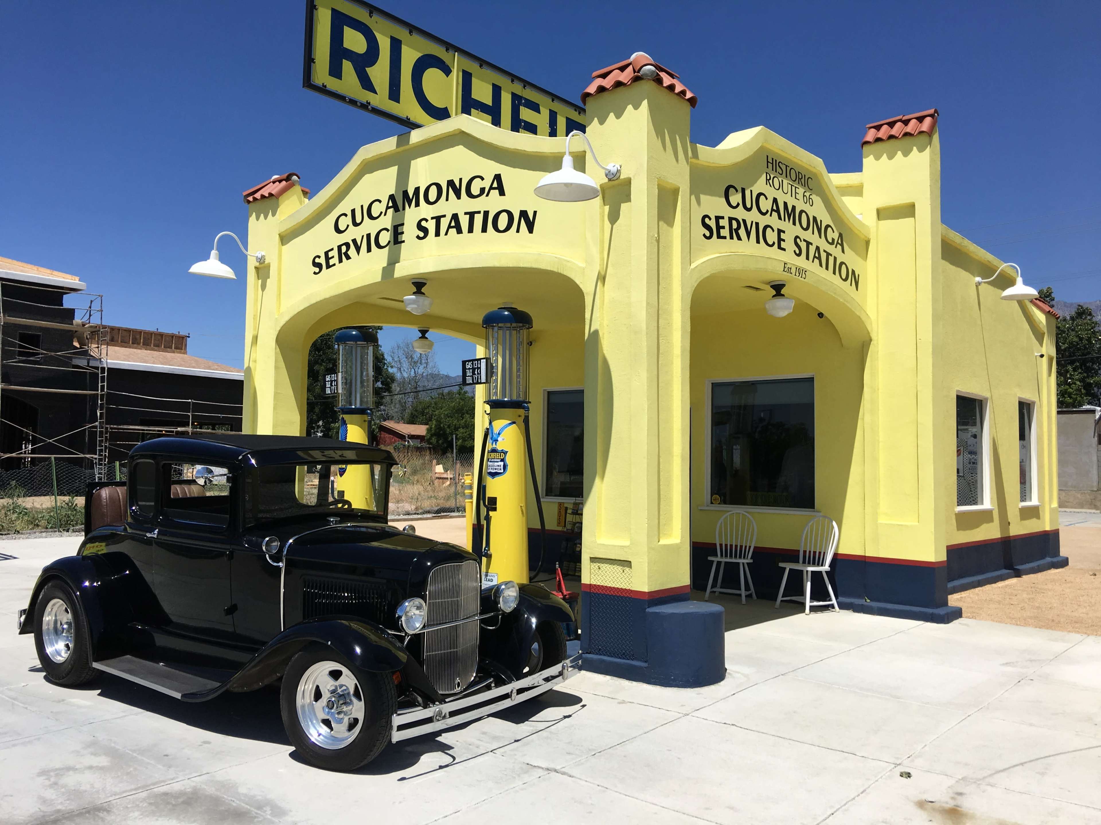 A vintage black car is parked in front of a bright yellow historic service station with the words "Cucamonga Service Station" on the awning.