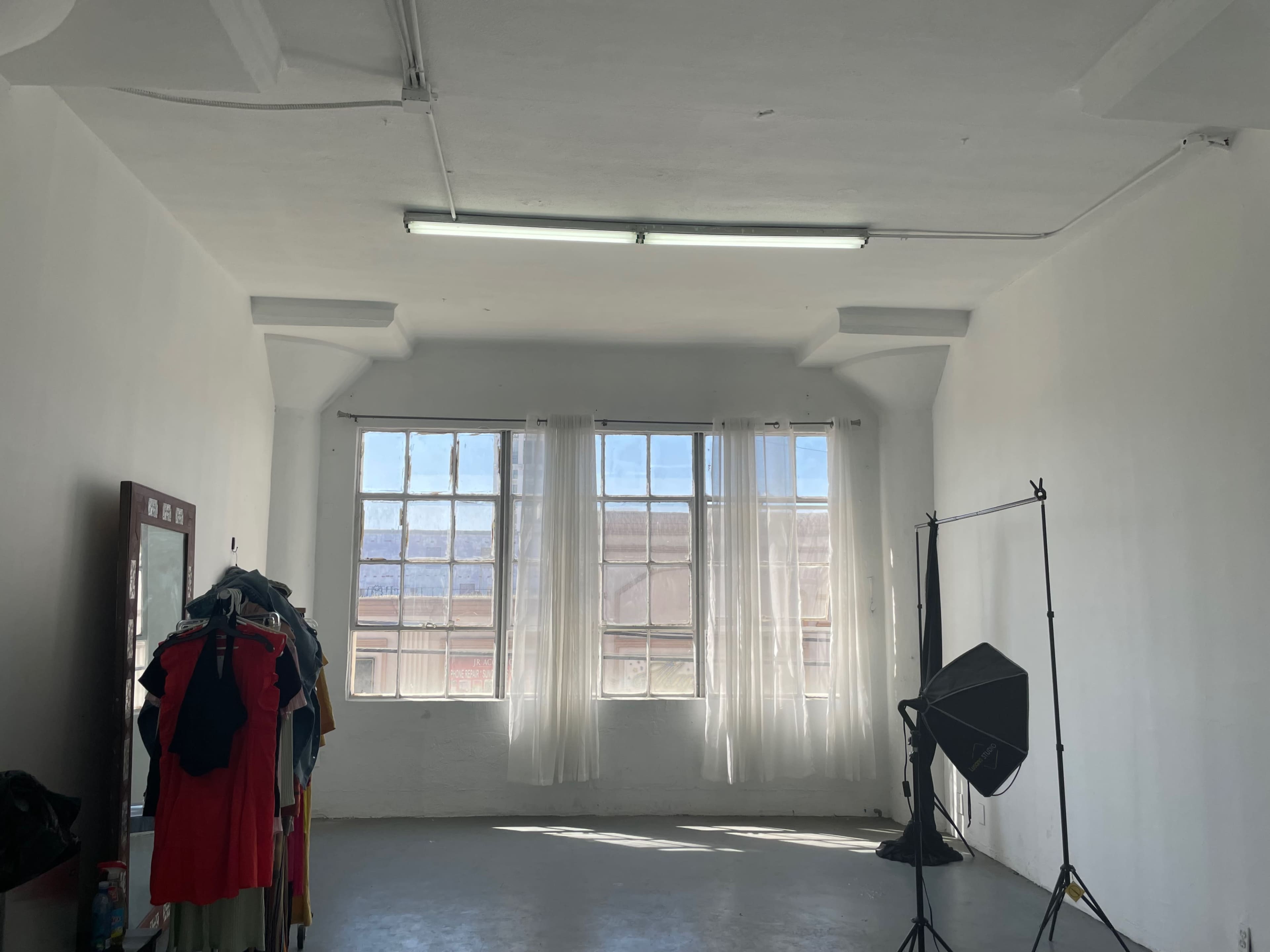 The image shows a bright, empty room with large windows, sheer white curtains, a mirror on one wall, a clothing rack, and a photography backdrop stand.