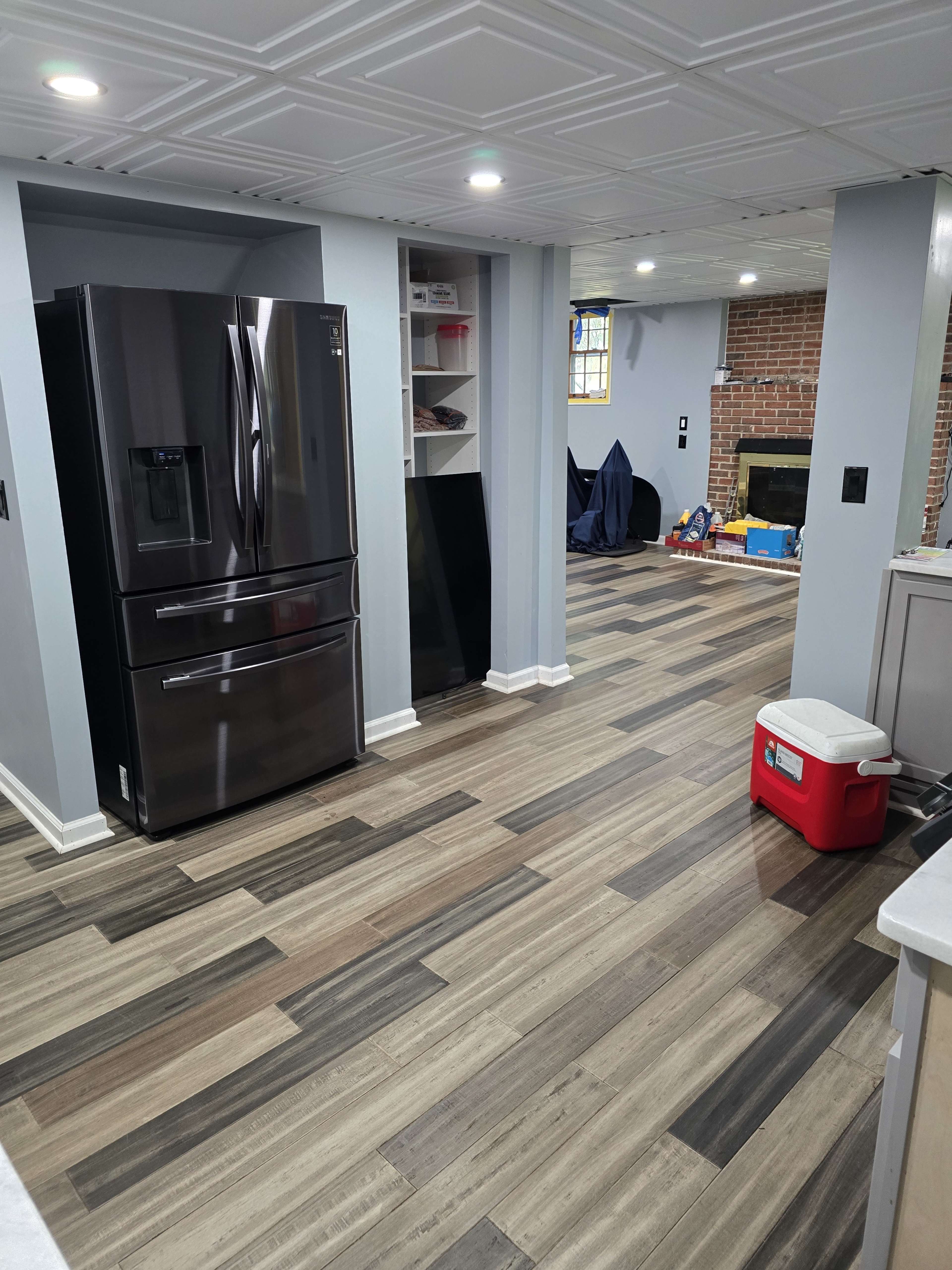 The image shows a modern kitchen with a black refrigerator, gray walls, and wooden flooring, featuring a red cooler and an open space leading to a living area with a fireplace.