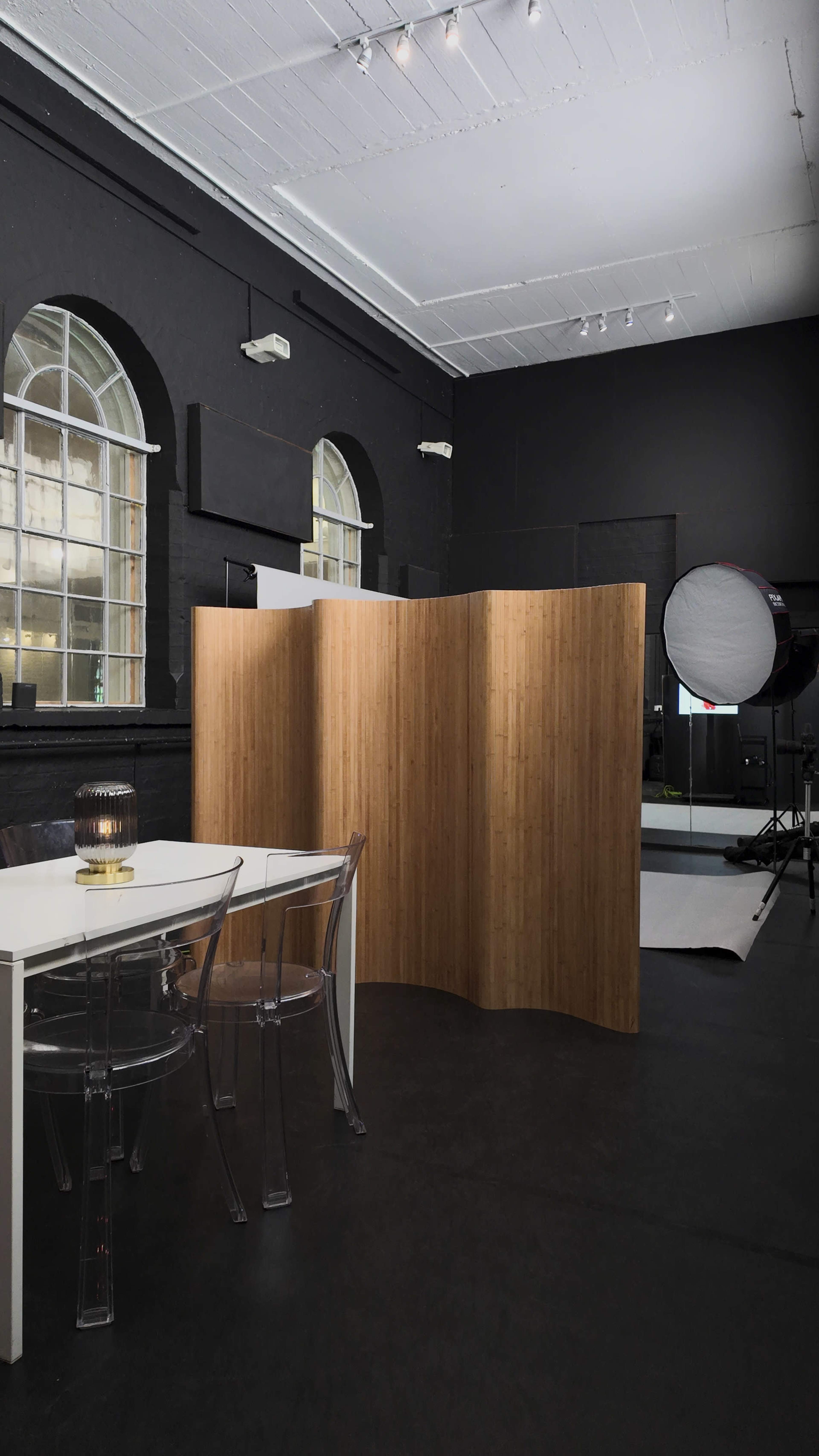 The image shows a modern studio space with black walls, a curved bamboo partition, a clear acrylic dining table with chairs, and professional lighting equipment in the background.