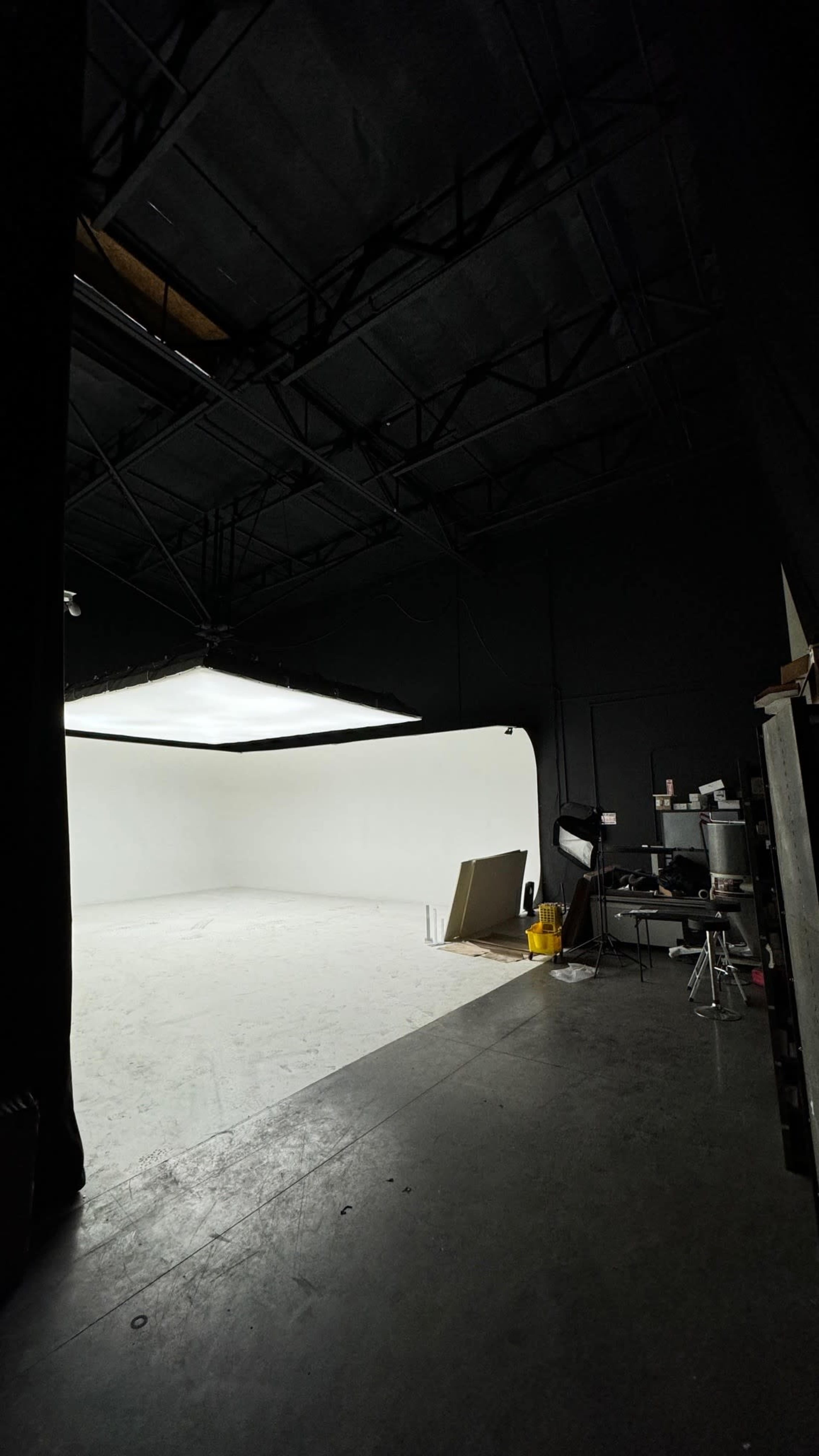 The image shows an empty studio space with a white backdrop and dim lighting, featuring a stool and some equipment on the side.