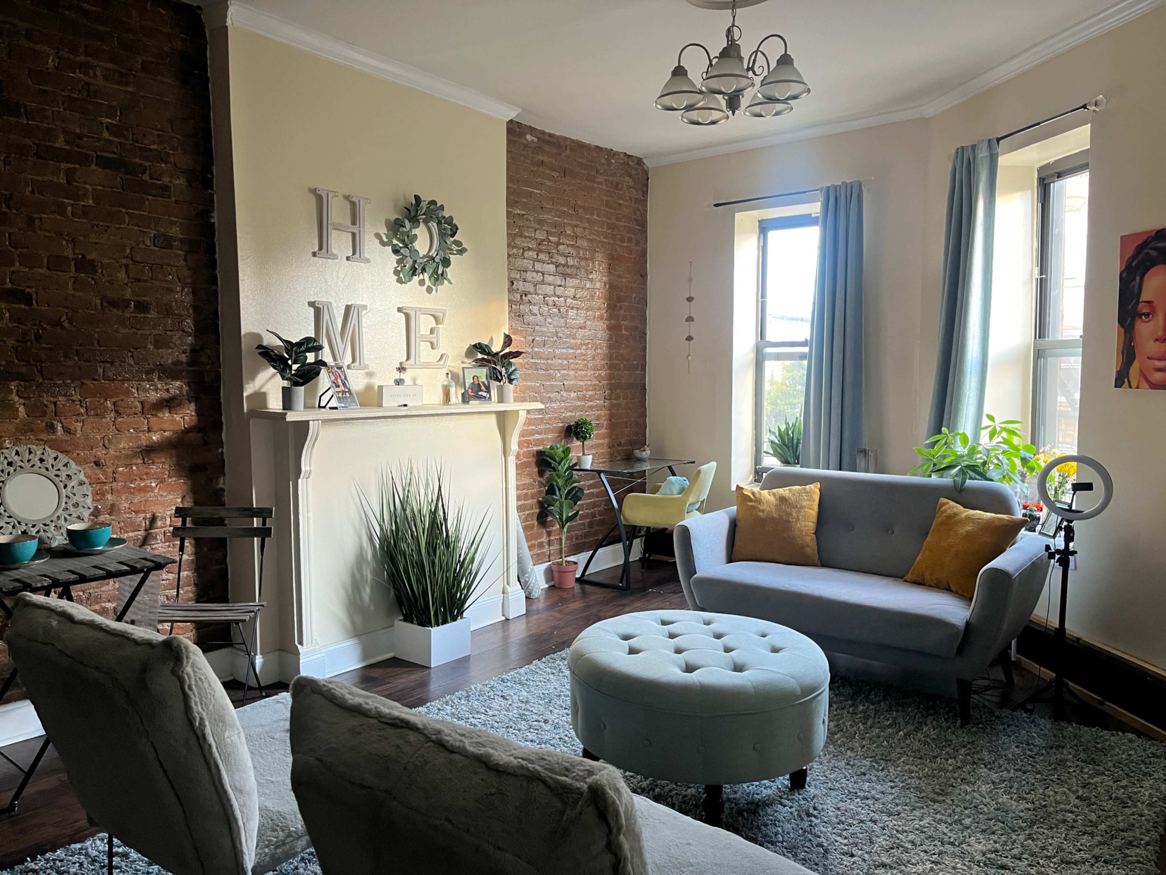 A cozy living room featuring a gray sofa, a round ottoman, and decorative plants against a backdrop of exposed brick walls.