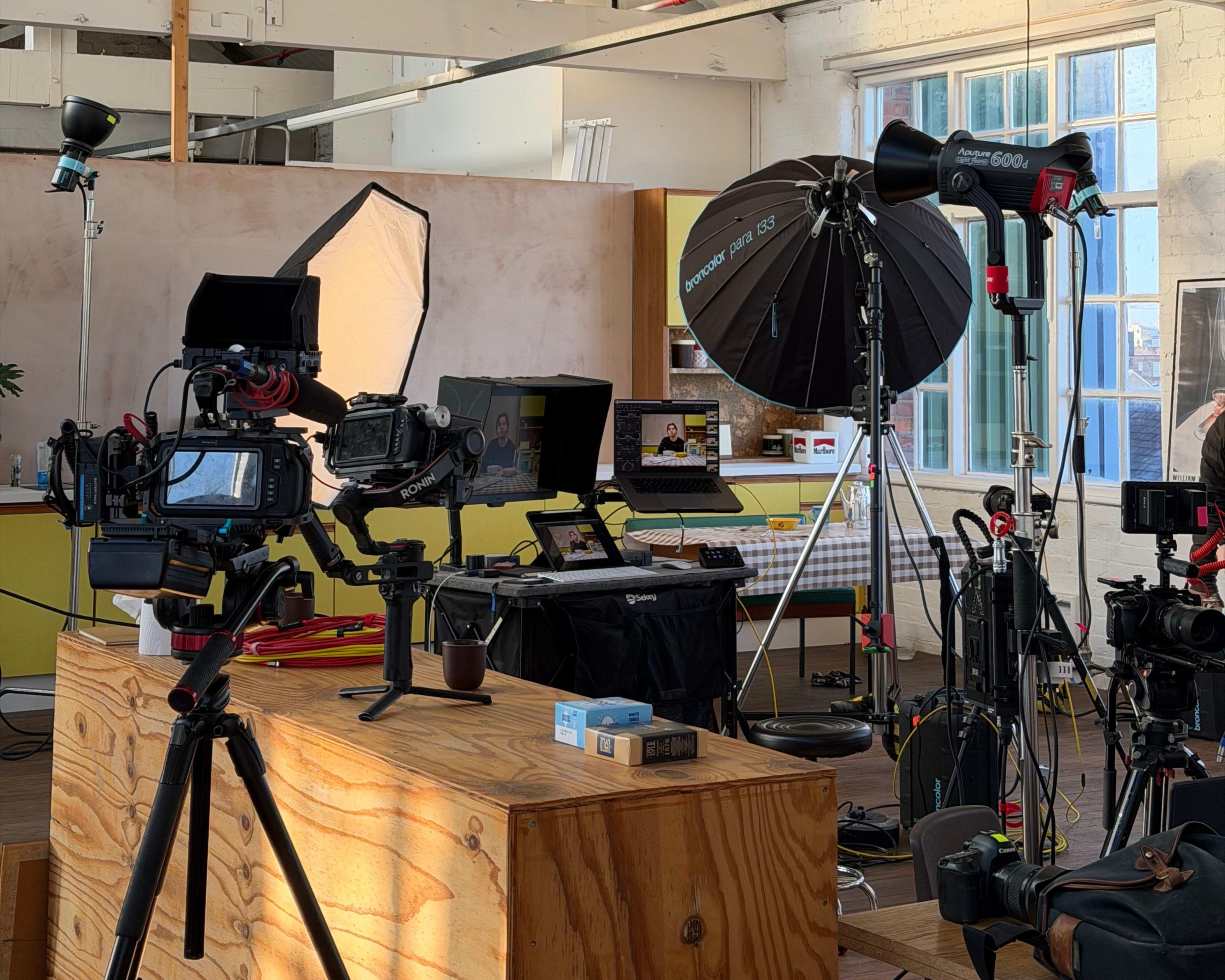 The image shows a photography studio setup with various cameras, lighting equipment, and a computer workstation.