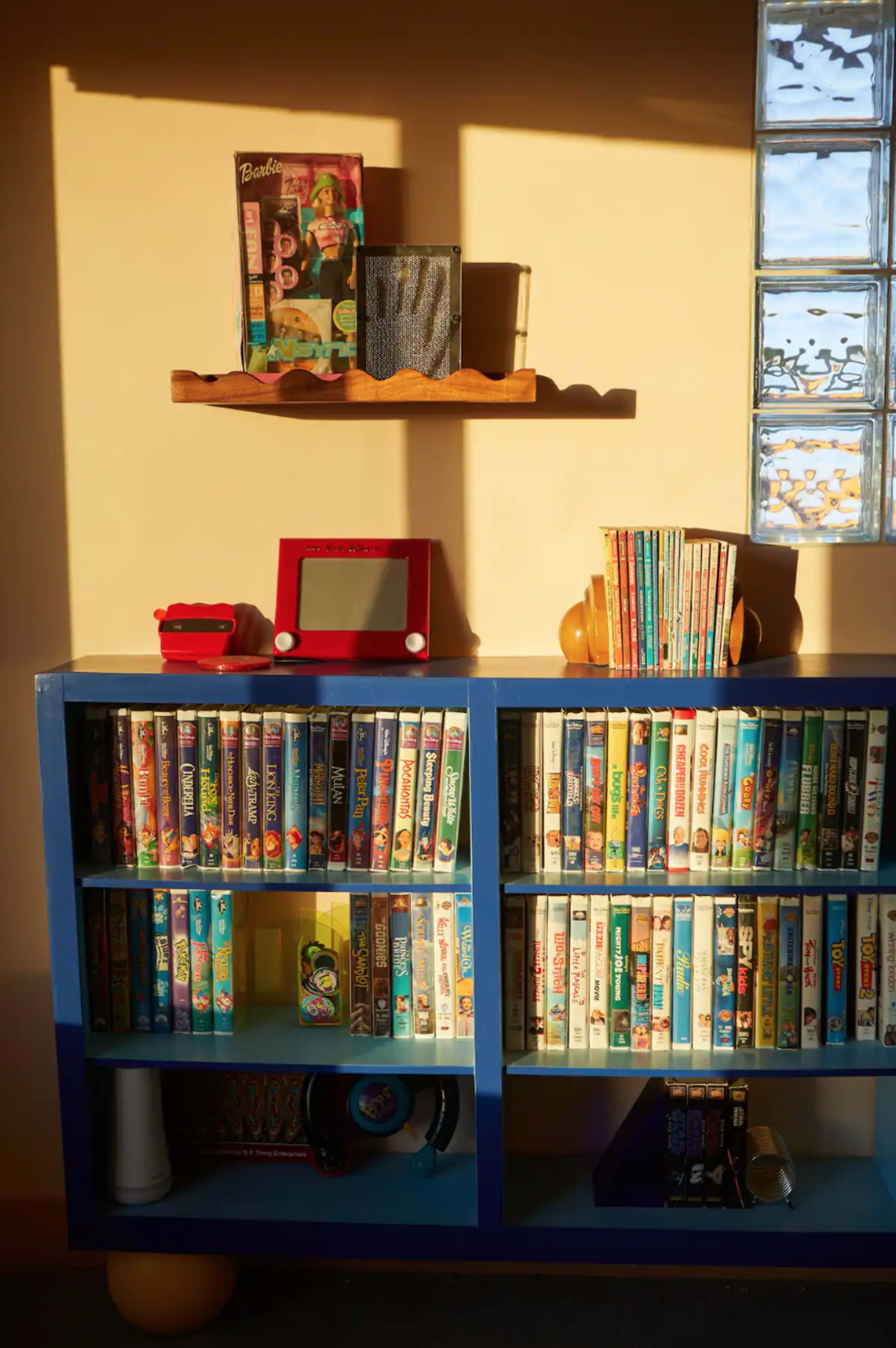 A bookshelf filled with various video cassettes, a small wooden shelf above displaying a few items, and sunlight illuminating the scene.