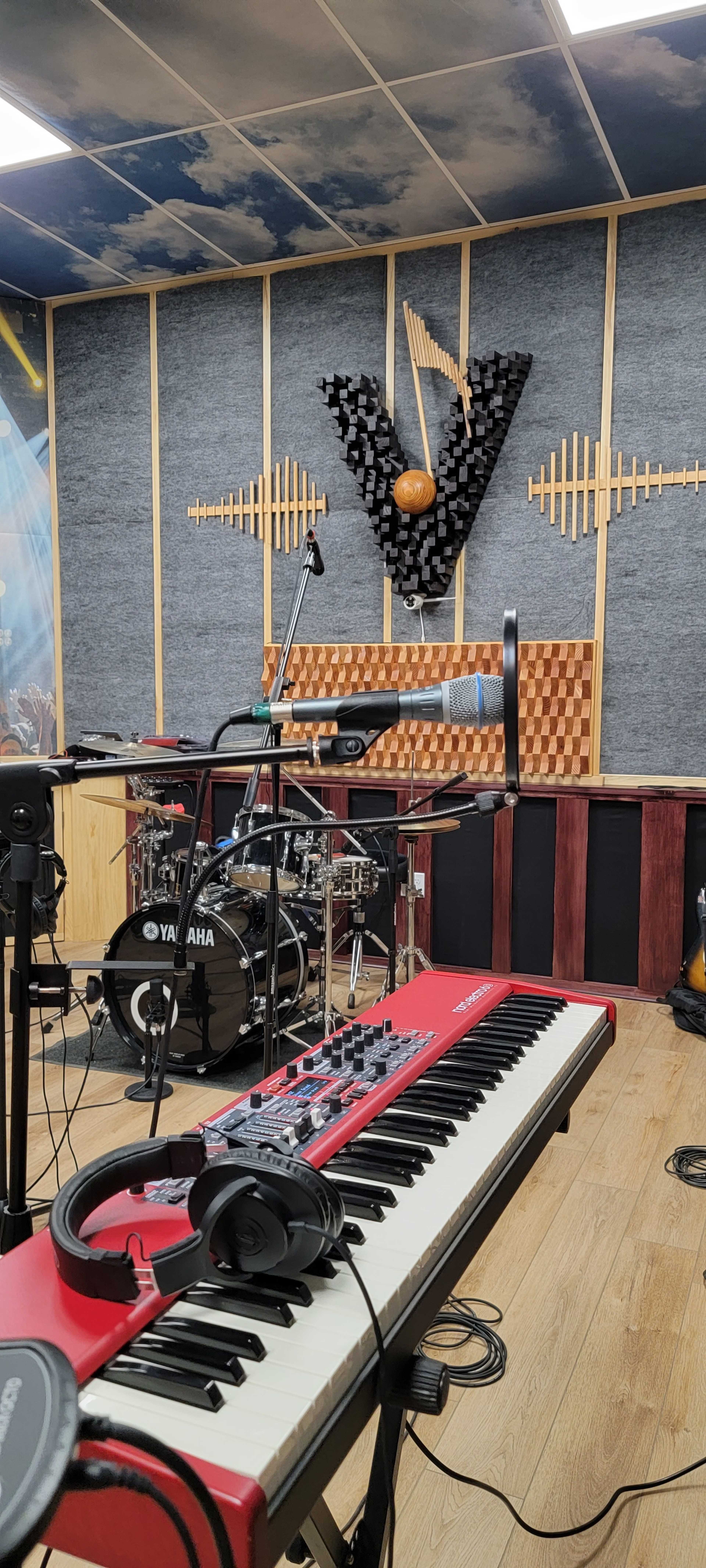 A music studio with a red keyboard, microphone, and a drum set arranged against a textured wall featuring wooden sound panels.