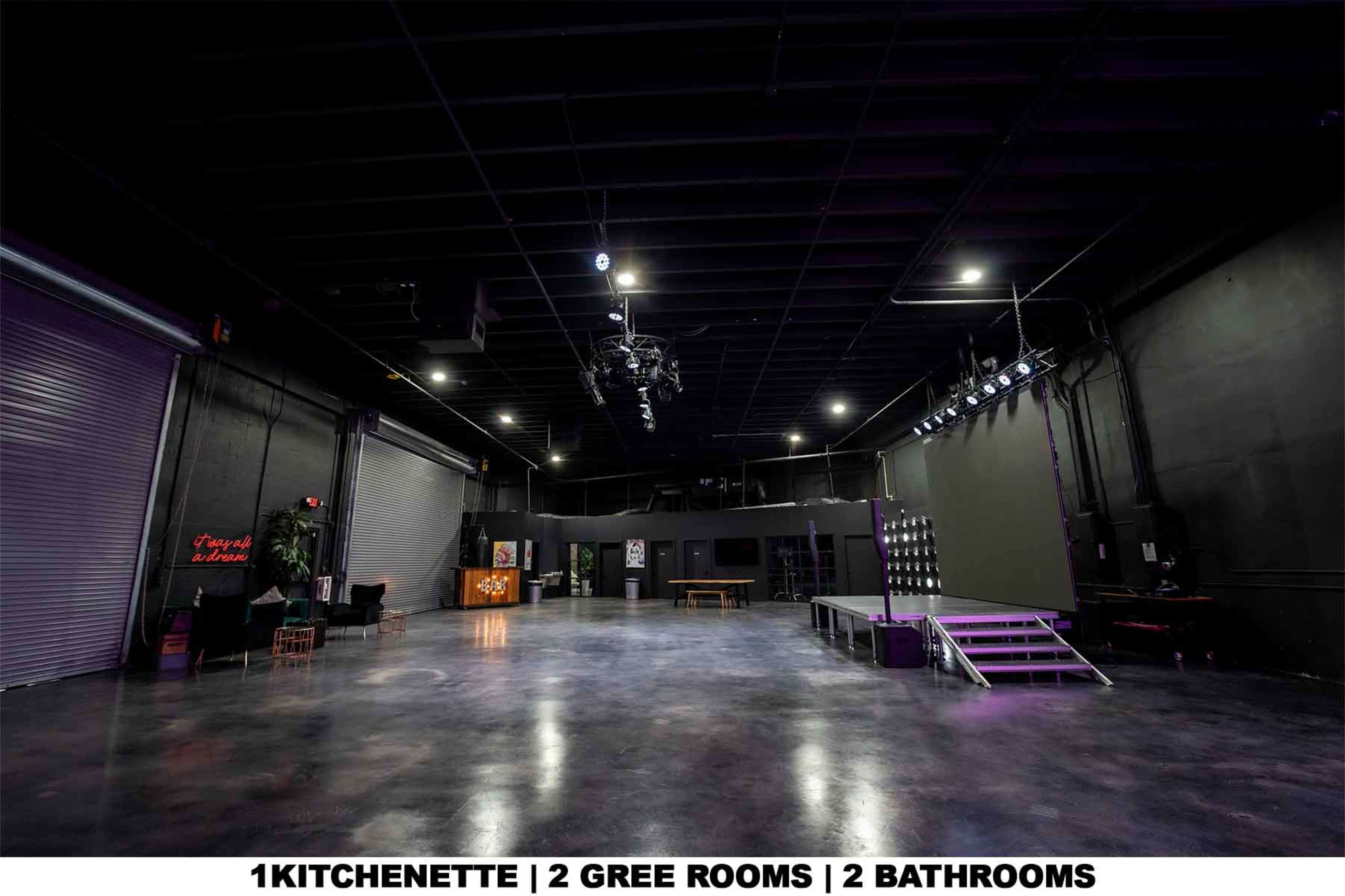 The image shows a spacious industrial studio or rehearsal space with a polished concrete floor, two large roll-up doors, and a small stage area illuminated by overhead lights.
