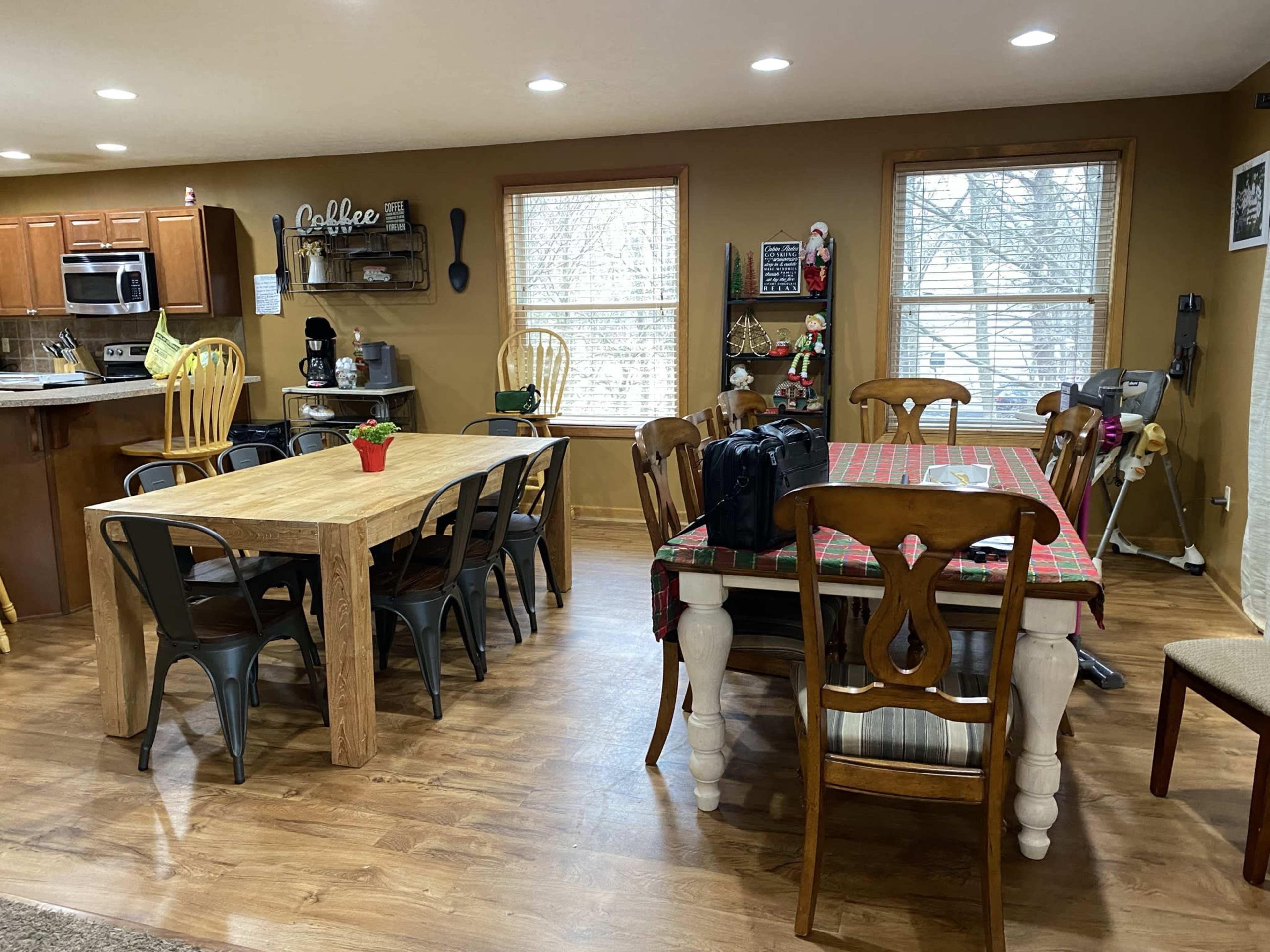 A spacious kitchen and dining area featuring a large wooden dining table, several chairs, and a kitchen counter in a well-lit room with windows.