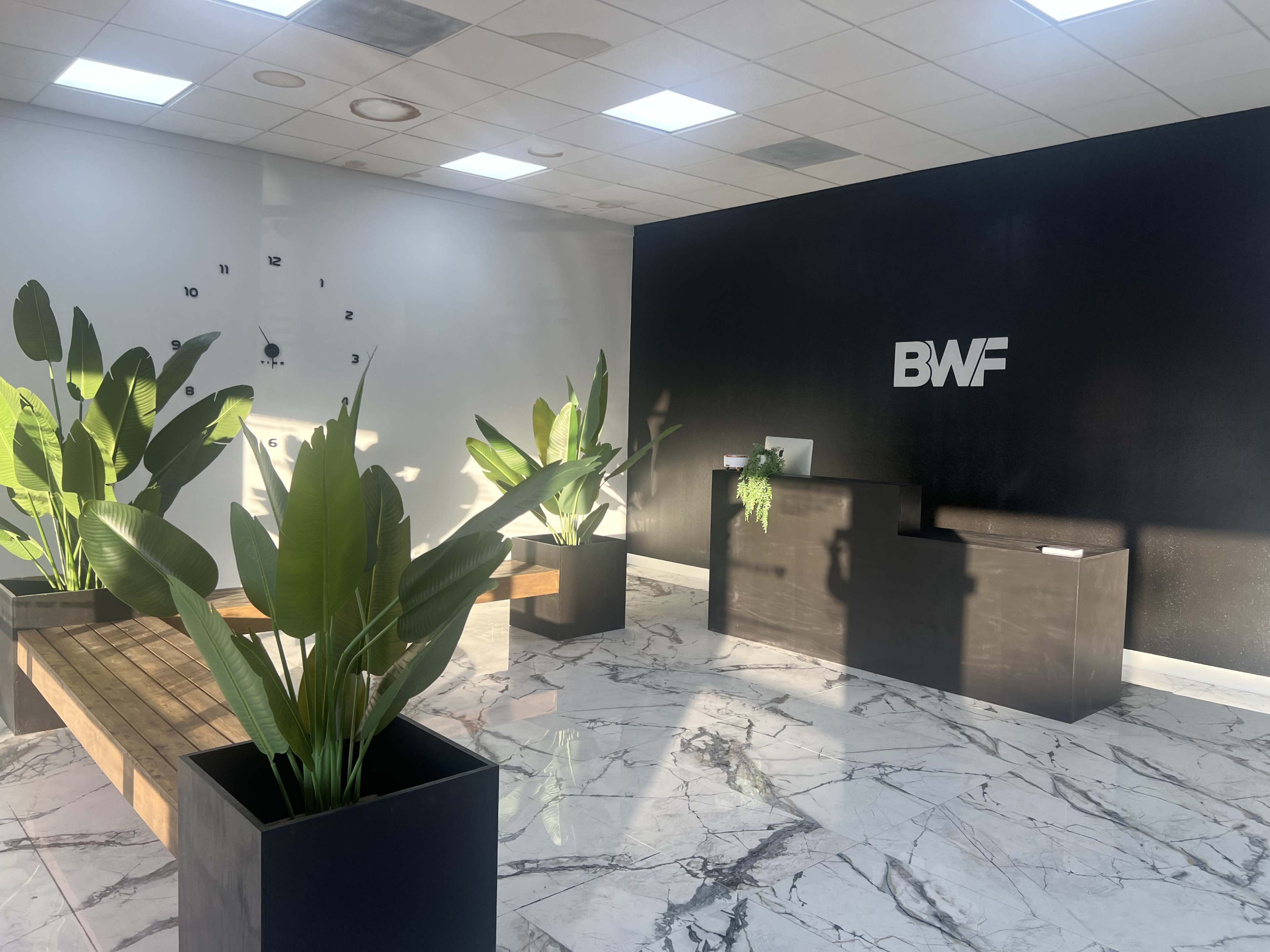 The image shows a modern reception area with a black wall featuring the letters "BWF," a wooden bench, and large plants in square pots on a marble floor.