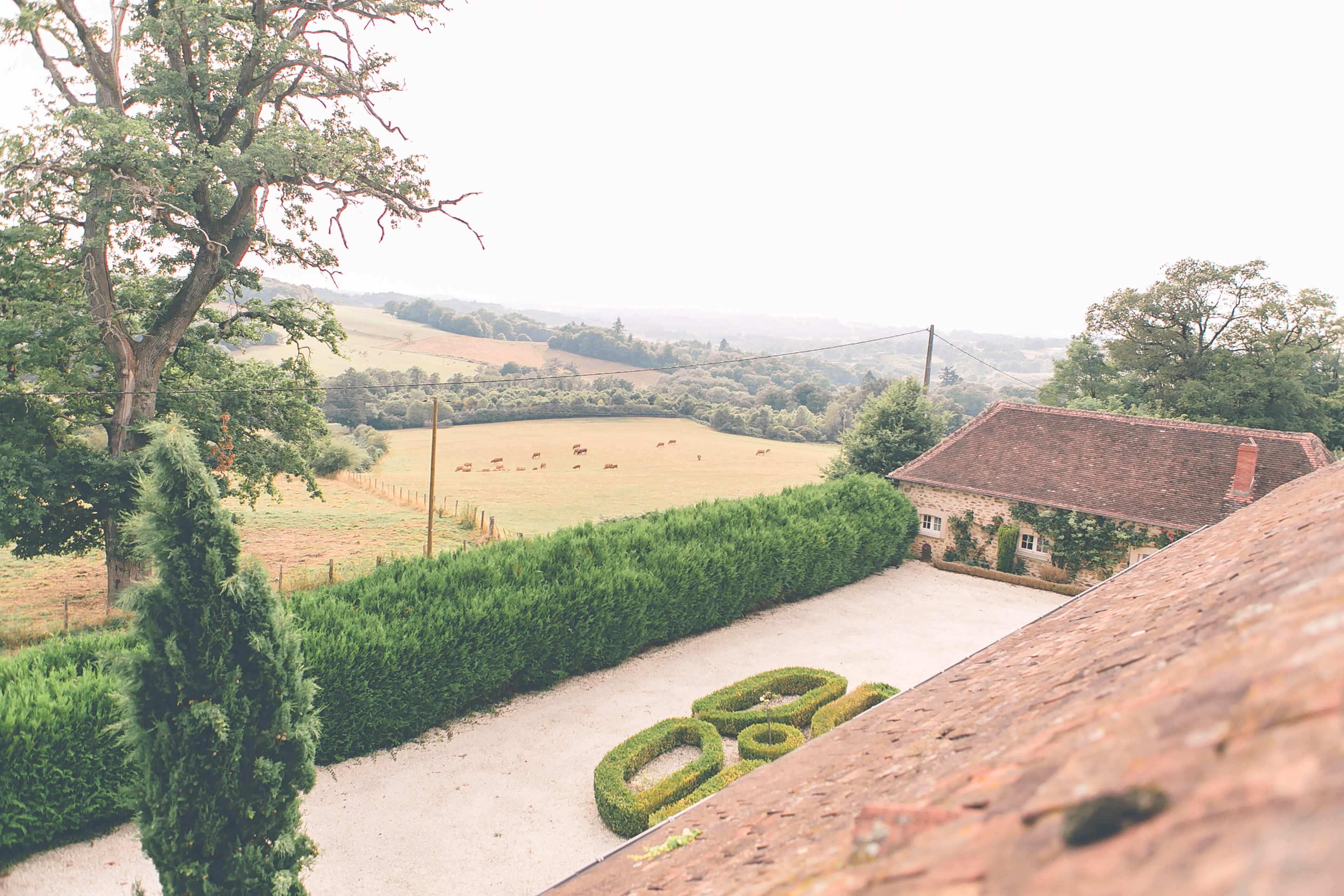 The image shows a rural landscape featuring a farmhouse surrounded by neatly trimmed hedges and fields with cattle in the distance.