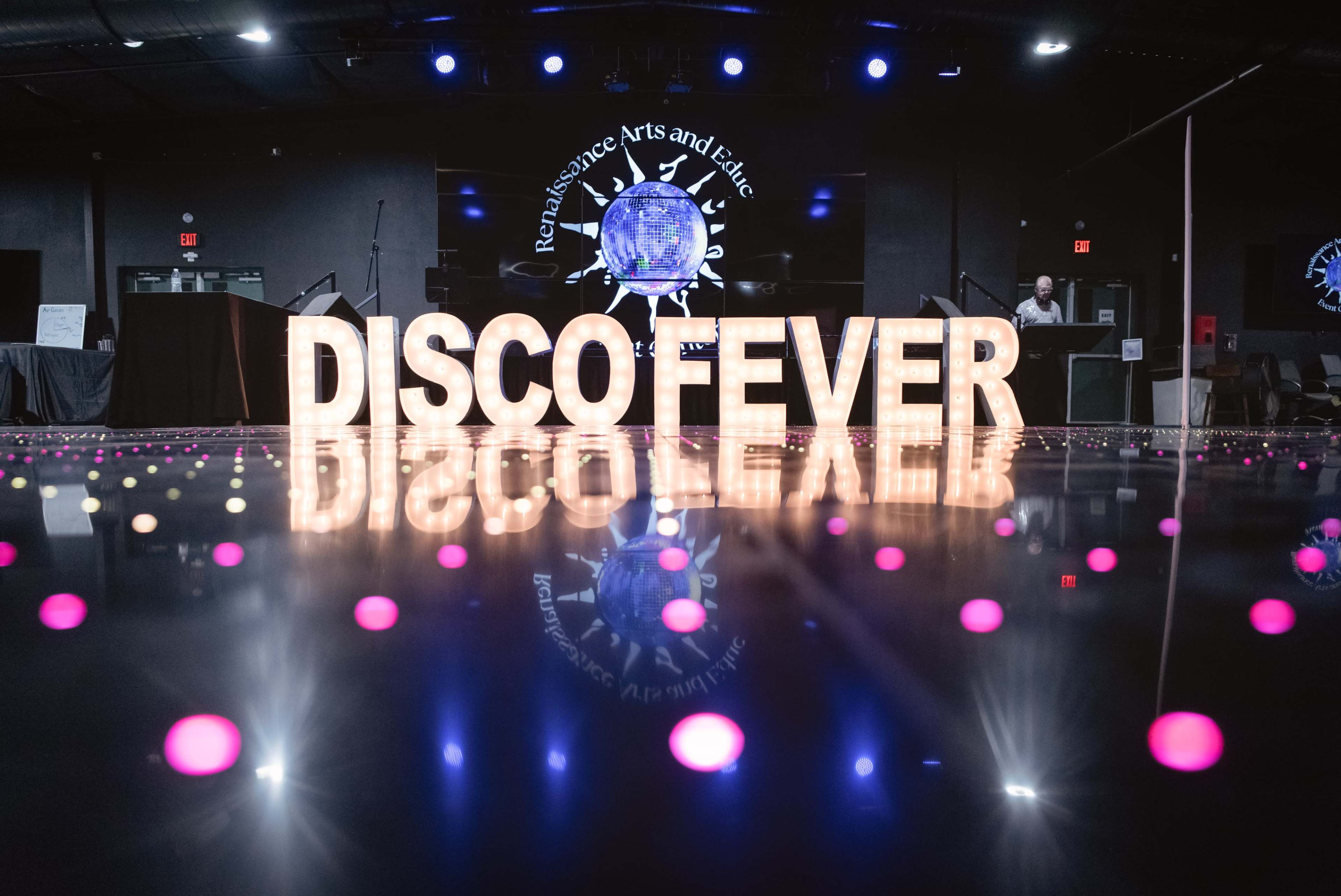 The image shows a dance floor with bright colored lights and a large illuminated sign reading "DISCO FEVER" against the backdrop of a venue decorated for an event.