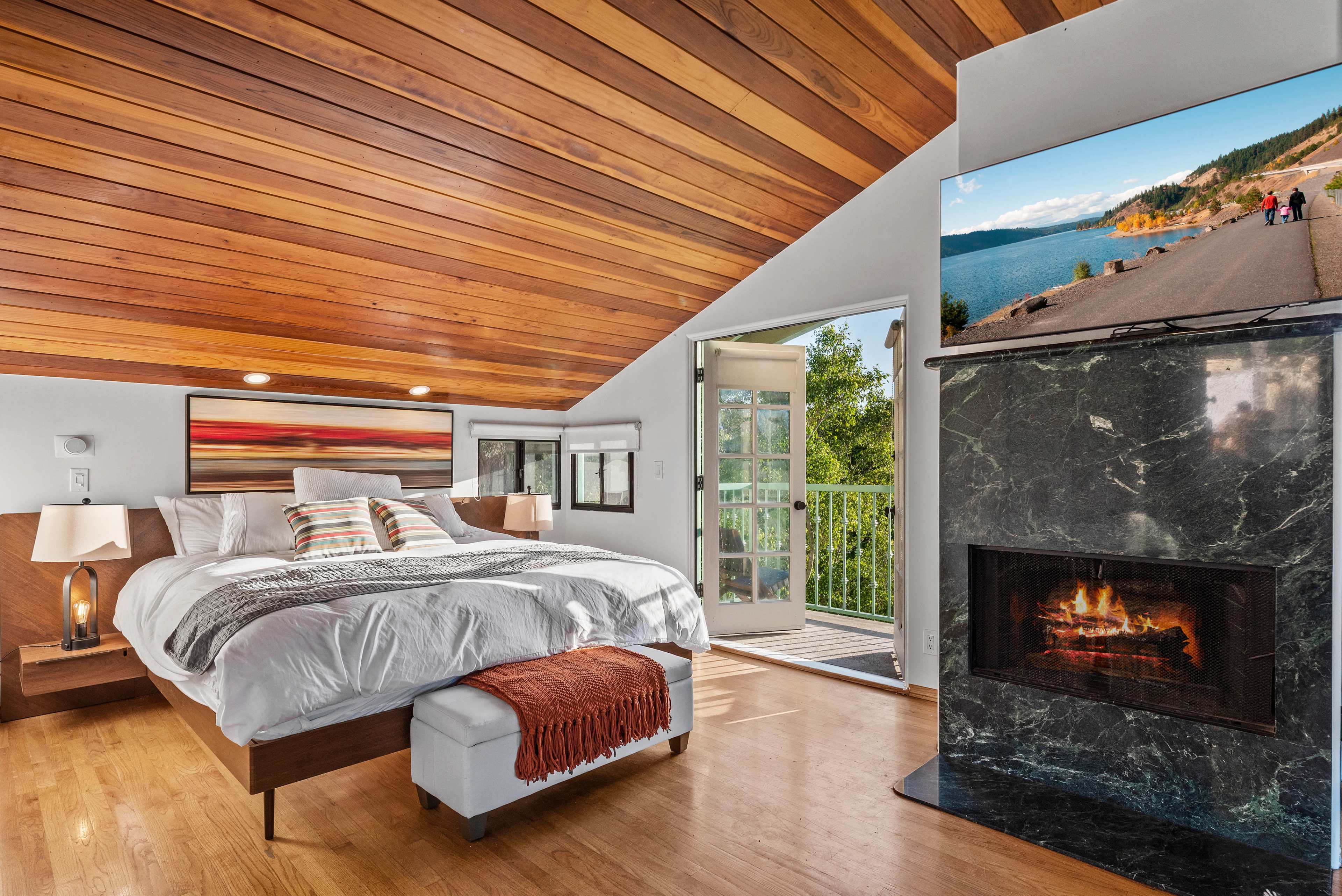 The image shows a modern bedroom with a wooden ceiling, a large bed with white linens, and a fireplace, opening onto a balcony with a view.