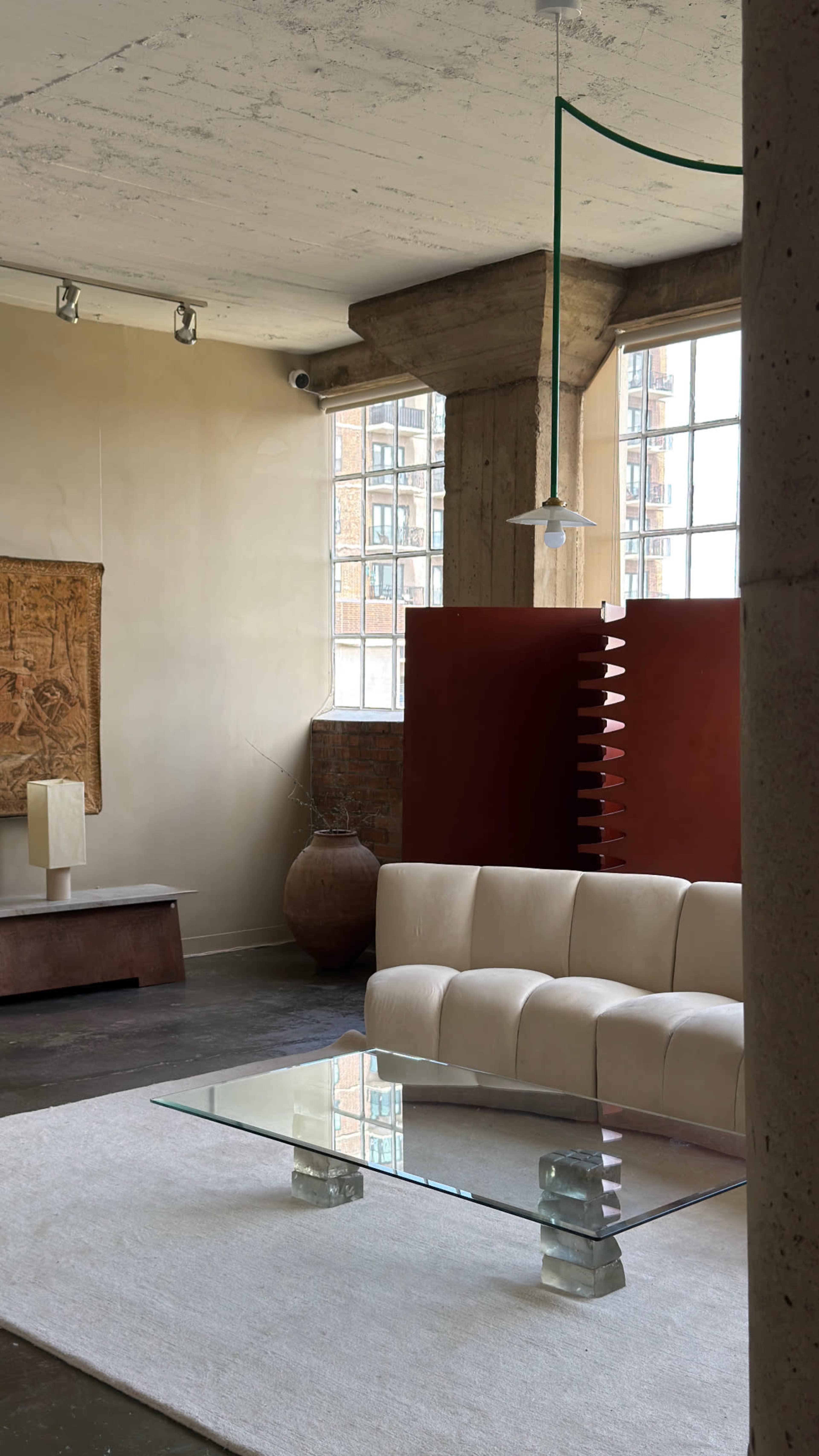 The image shows a minimalist interior space featuring a cream-colored sofa, a glass coffee table, and large windows allowing natural light to enter.