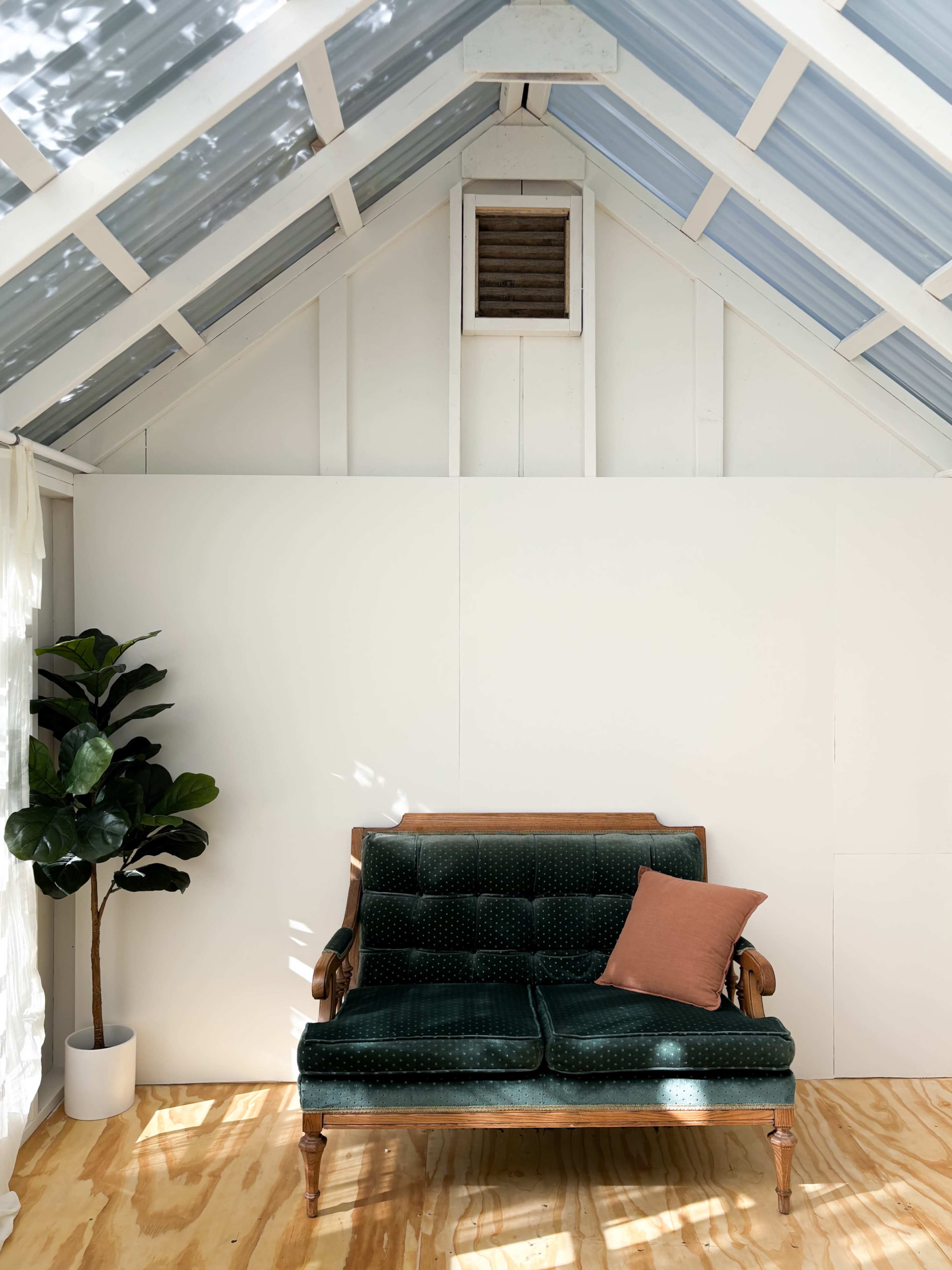 The image shows a bright, minimalistic indoor space featuring a green upholstered loveseat with a lone orange cushion, a potted plant, and wooden flooring beneath a slanted roof with blue panels.